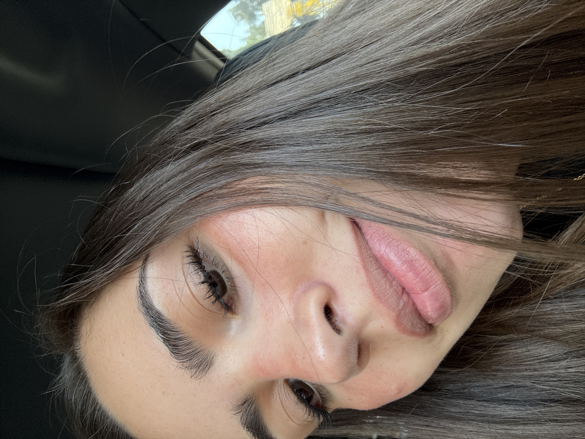 A close-up, angled portrait shows a young woman with light skin and dark brown hair, partially in focus. Her mouth is slightly open, with her tongue partially visible. She has dark eyebrows and eyelashes, and her eyes are brown. The background appears to be the interior of a car with a glimpse of trees visible through the window. There is no visible evidence to suggest the location is the city of Puebla, Mexico.