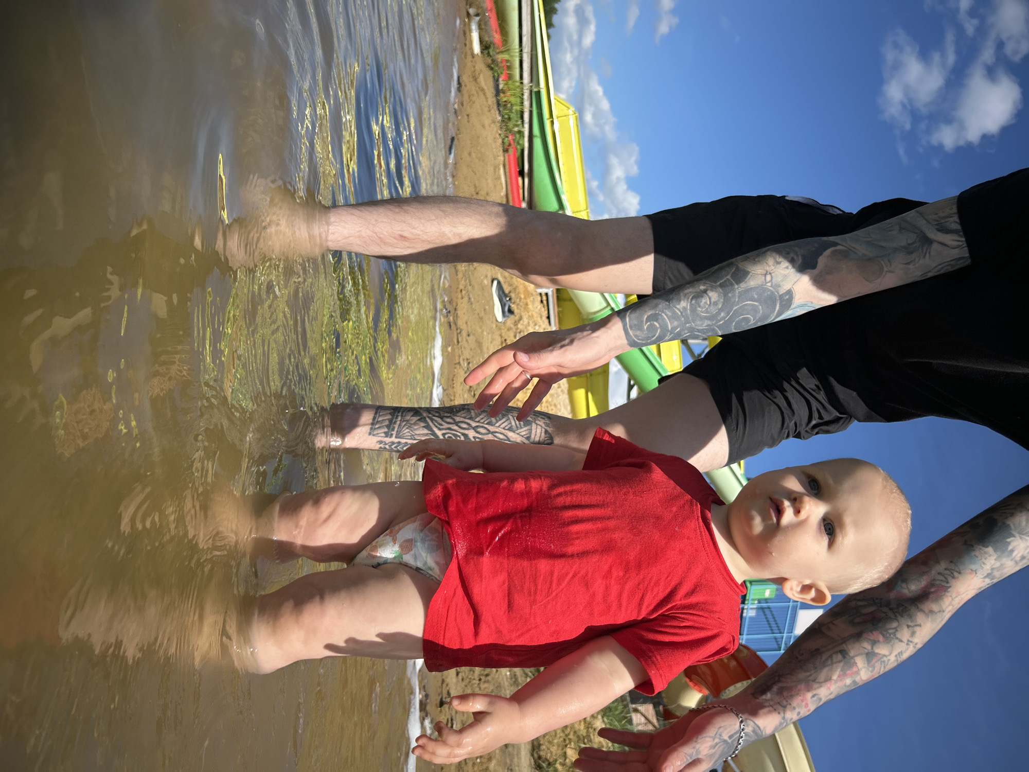 The image depicts a toddler standing in shallow, murky water. The toddler is wearing a red t-shirt and a diaper with a floral pattern. Behind the toddler, the legs of an adult are visible, one leg with extensive tattoos. The adult's hand is reaching towards the toddler. In the background, there are colorful water slides, a sandy shore, and a blue sky with white clouds. The location is identified as the city of Yubileynyy, Russia. The overall scene suggests a recreational setting, possibly a water park or a lakeside area on a sunny day.