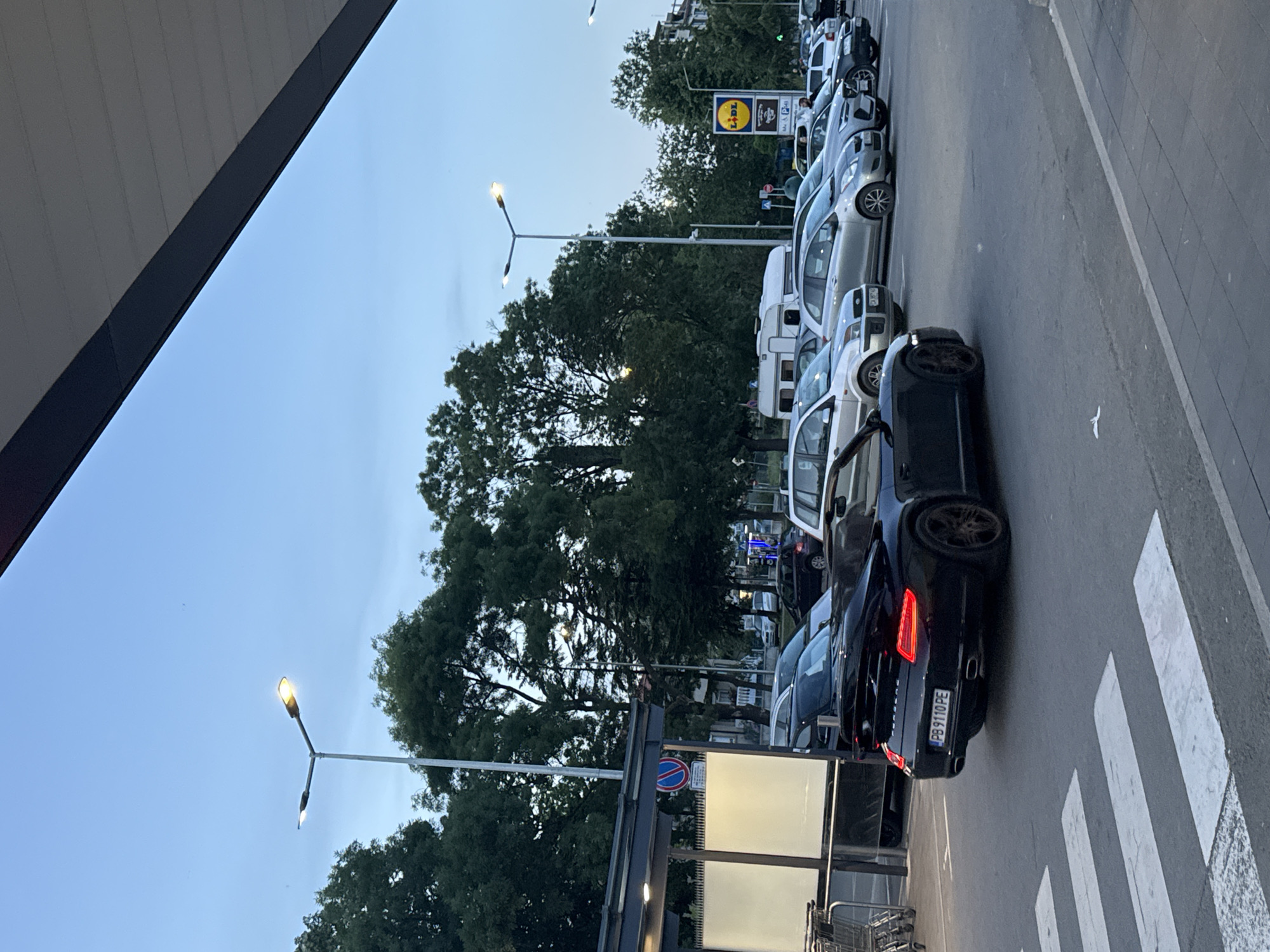A black Porsche convertible with license plate PB 9110 PE is parked in a lot, its brake lights illuminated. The car is positioned on a crosswalk in front of a covered structure with white rectangular signs. Other cars, including silver sedans, a white RV trailer, and a black SUV, are visible in the background, parked in a lot. A LIDL sign is displayed prominently. The scene is set against a backdrop of trees and a clear, overcast sky, suggesting it is dusk. The environment appears to be a commercial area, possibly a parking lot of a supermarket. The location is identified as Tsarevo, Bulgaria. There are no visible individuals interacting with the cars or the environment.