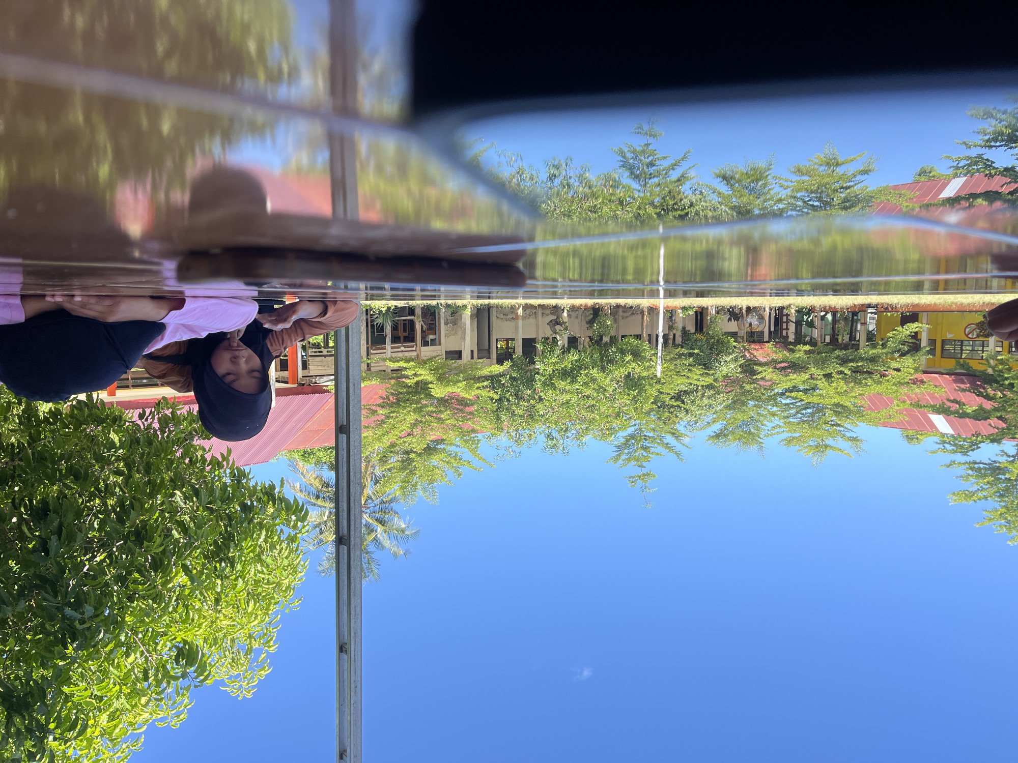 Two young females are lying on a reflective, brown, wooden table outdoors. One female, wearing a black hijab and brown long-sleeved shirt, is propped up on her elbows and looking at a smartphone in her hands. The other female, wearing a black hijab and pink long-sleeved shirt, is lying on her stomach with her head resting on her arms, facing away from the camera. A dark-colored object, possibly a smartphone, is positioned horizontally on the table in the foreground.
The background features a row of low-rise buildings with red roofs, a line of green trees, and a clear blue sky. A thin, metallic pole stands vertically on the right side of the image. The scene appears to be a school or public area. The reflection on the table shows the buildings, trees, and sky. The overall impression is one of a sunny, daytime setting in a tropical or sub-tropical environment, possibly a school campus in Pompanua, Indonesia, given the architectural style and vegetation.