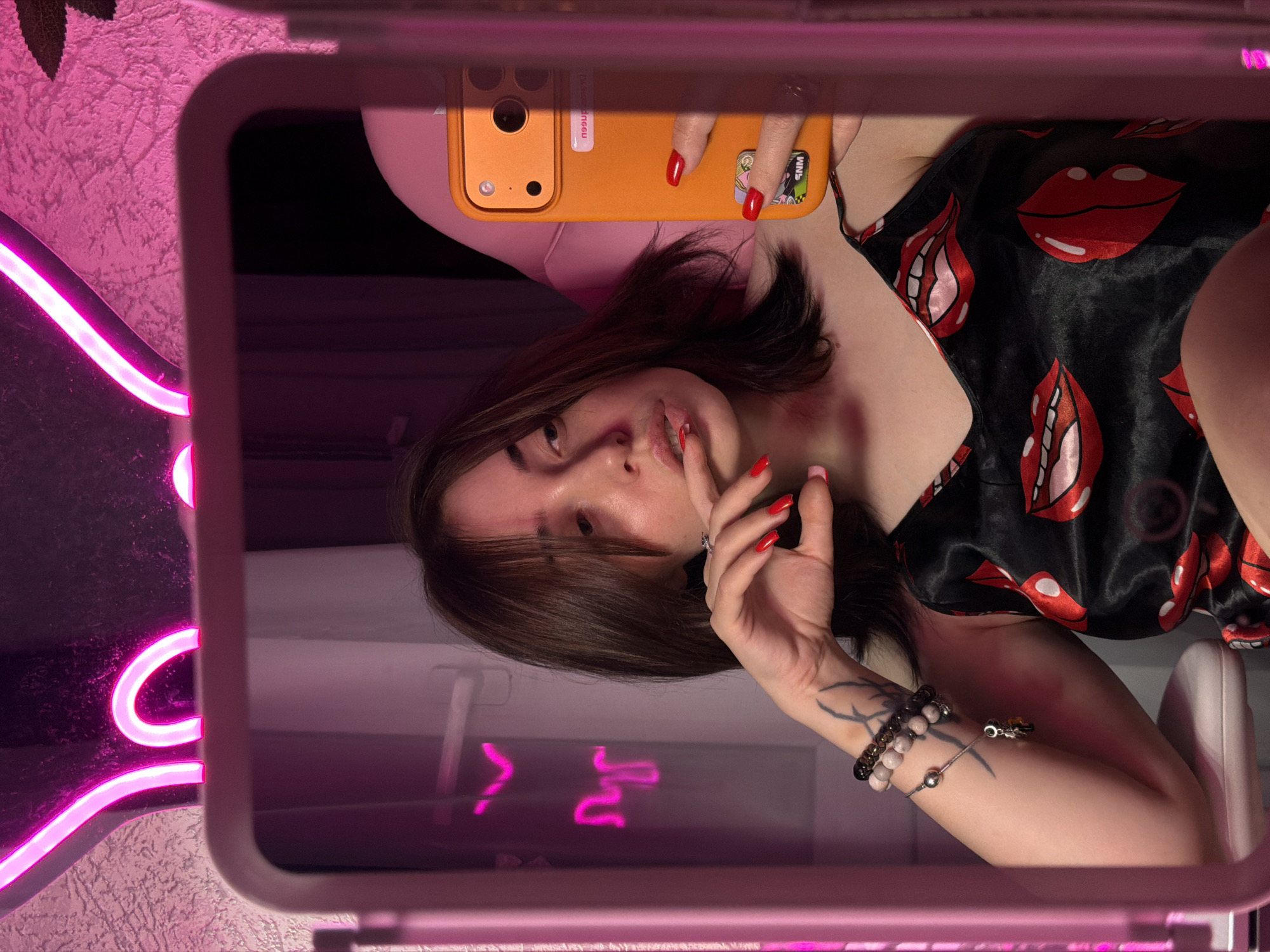 A young woman with dark hair and fair skin is reflected in a mirror. She is wearing a black satin slip with a pattern of red lips. She is holding an orange smartphone in her right hand, taking a selfie. Her nails are painted bright red. A neon pink light fixture, shaped like rabbit ears, is visible above the mirror. The background is a textured wall. A pink gaming chair is partially visible. A tattoo is visible on her left arm, as are several bracelets.
