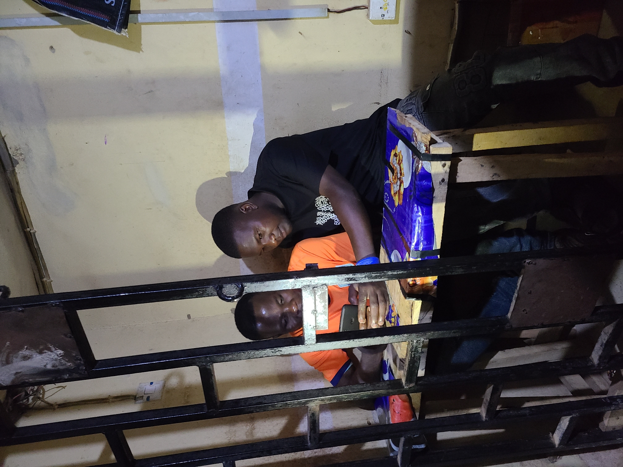 The image, taken with a vertical orientation, captures two men in what appears to be an indoor or semi-enclosed, dimly lit space. The overall setting suggests a utilitarian environment with exposed wiring and simple structures.
In the foreground, partially obscured by black metal railings, a man with dark skin is visible. He wears an orange t-shirt and is focused on a black smartphone held in his hands. His expression is neutral and concentrated. An orange sneaker is visible on his left foot.
Above and slightly behind this man, another man with dark skin is positioned. He wears a black t-shirt and looks directly towards the viewer with a serious or neutral expression. He appears to be leaning over a wooden structure.
The immediate background consists of light yellow or beige walls, which show some signs of wear and dirt. On the upper part of the wall, an electrical outlet with a brown wire extending from it is visible. Another electrical outlet can be seen through the metal railings on the lower left. A blue plastic covering with indistinct graphics, possibly packaging for a product, is wrapped around a wooden plank near the upper man. Further details of the background include dark wooden planks, forming shelves or part of a bed frame, and other dark areas. A small portion of a poster or sign with the letter "S" is visible in the top left corner of the frame.
The metal railings form a grid pattern, creating a barrier or structure within the space, possibly indicating a staircase, a bunk bed, or a mezzanine level. The dim lighting suggests it might be nighttime or an interior space with insufficient illumination. No specific weather conditions are discernible, and no strong emotions are evident from the subjects.