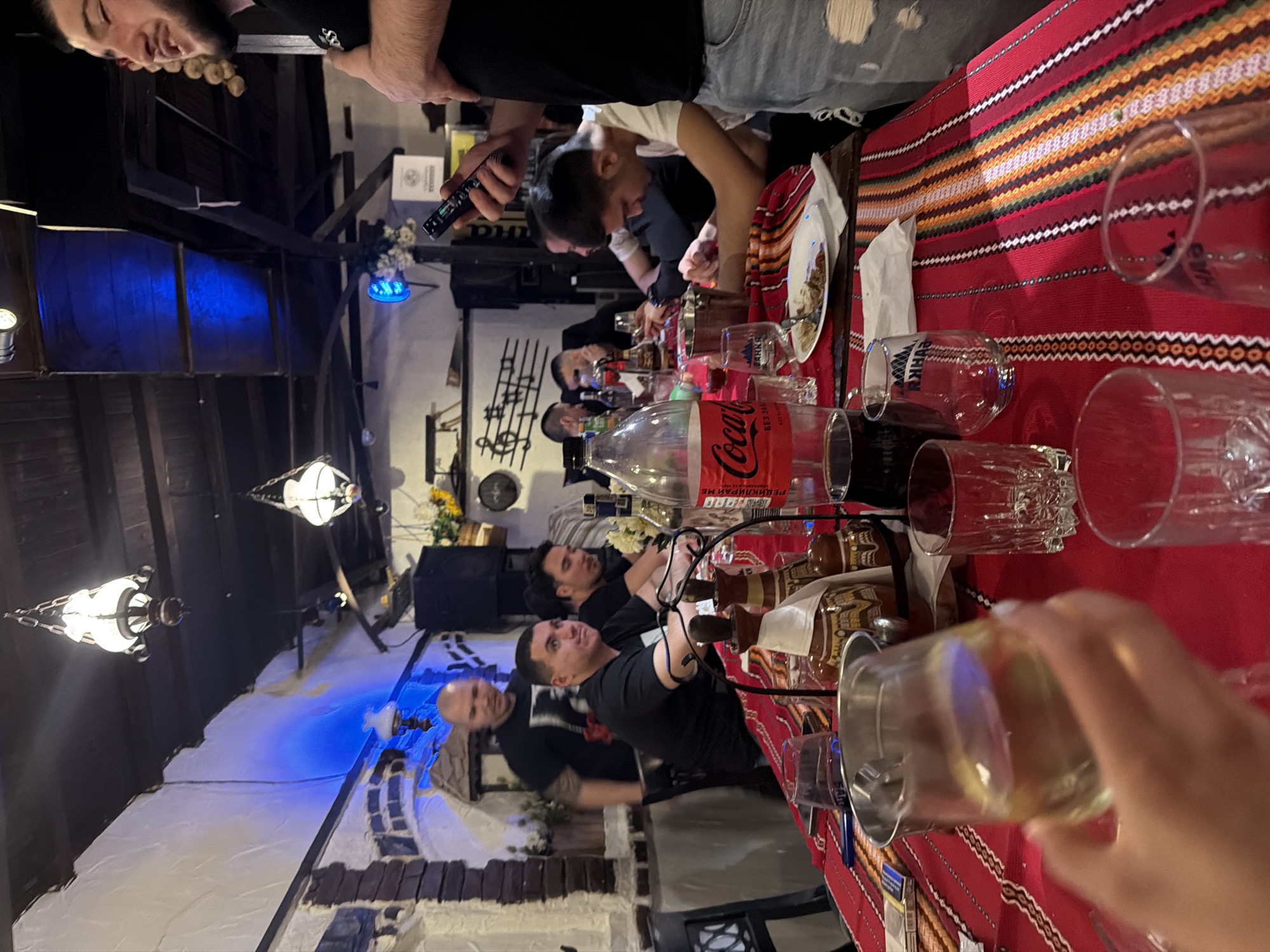 A casual gathering is taking place indoors at a restaurant with Bulgarian decor in Varna, Bulgaria. Multiple people sit around a long table covered with a red and patterned tablecloth. Beverages, including Coca-Cola and possibly alcoholic drinks, are visible on the table alongside food plates. The setting is informal, with hanging light fixtures contributing to the ambiance. People are engaged in eating, drinking, and socializing. The interior design features brickwork and blue lighting elements. The overall atmosphere suggests a relaxed and convivial dining experience.