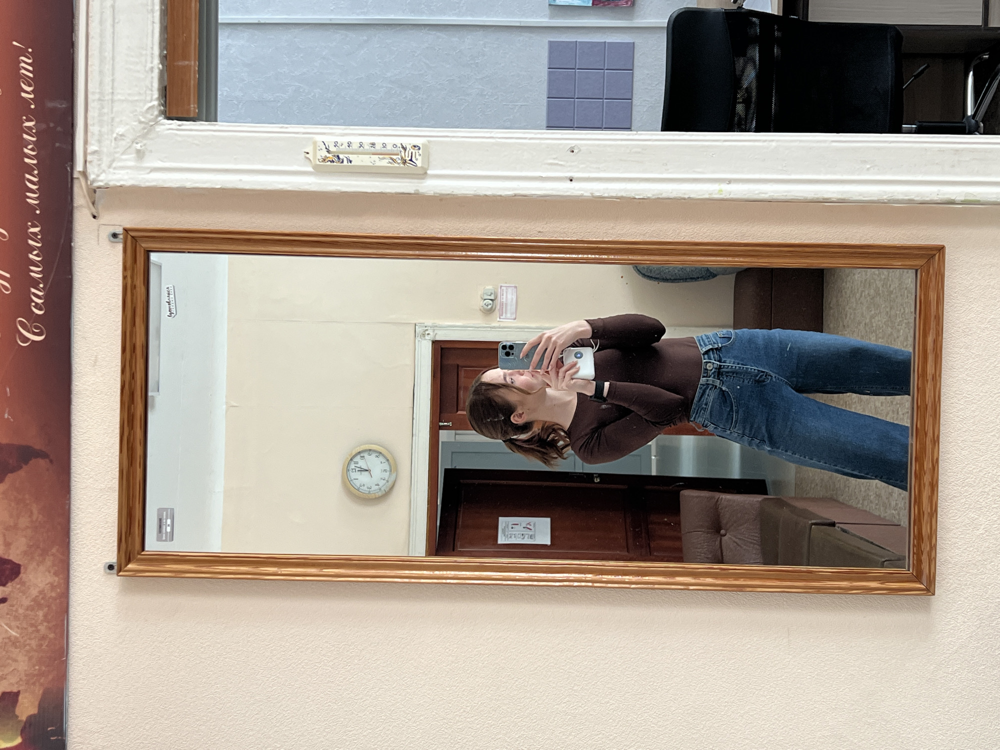 A mirror mounted on a peach-colored wall reflects a woman taking a selfie. The woman is wearing a brown long-sleeved shirt and blue jeans. The reflection shows a room with a wall clock, a closed door with a sign on it, and brown furniture. Through a window above the mirror, part of a room is visible, including a chair and a panel of square tiles. To the left of the mirror, a poster is partially visible. The location is likely in Chaikovsky, Russia, based on the text on the poster, "С самых малых лет!"
