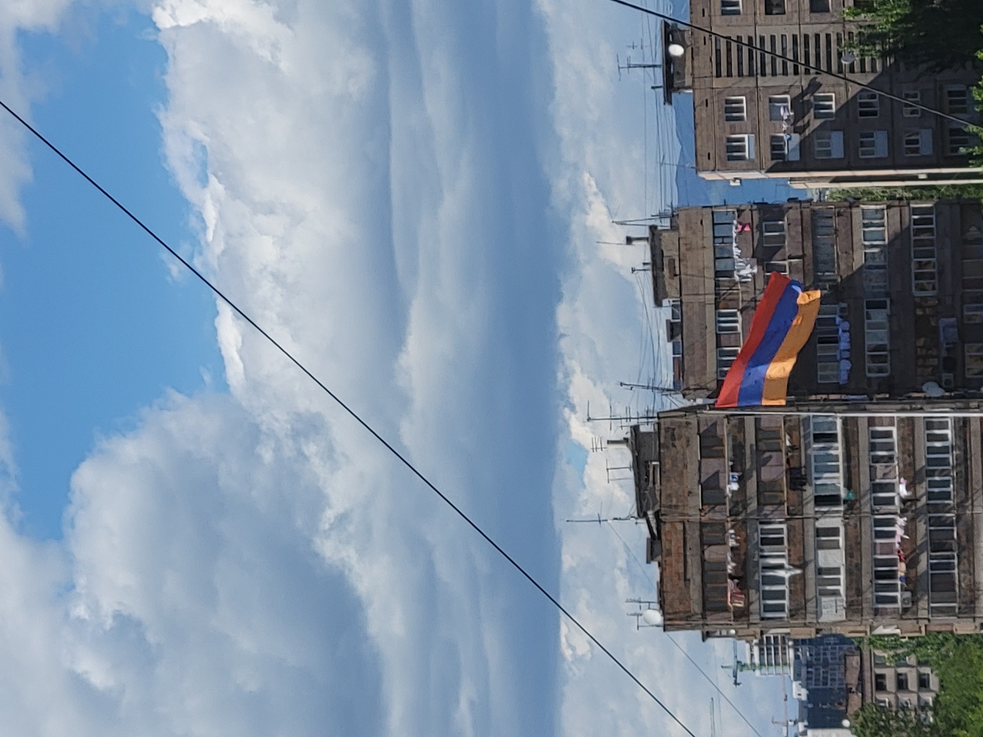 A vertical shot shows several tall concrete buildings in Yerevan, Armenia, against a partly cloudy sky. An Armenian flag hangs from one of the buildings. The buildings appear aged, and telephone/electrical wires span across the sky. Green foliage is visible near the base of the buildings.