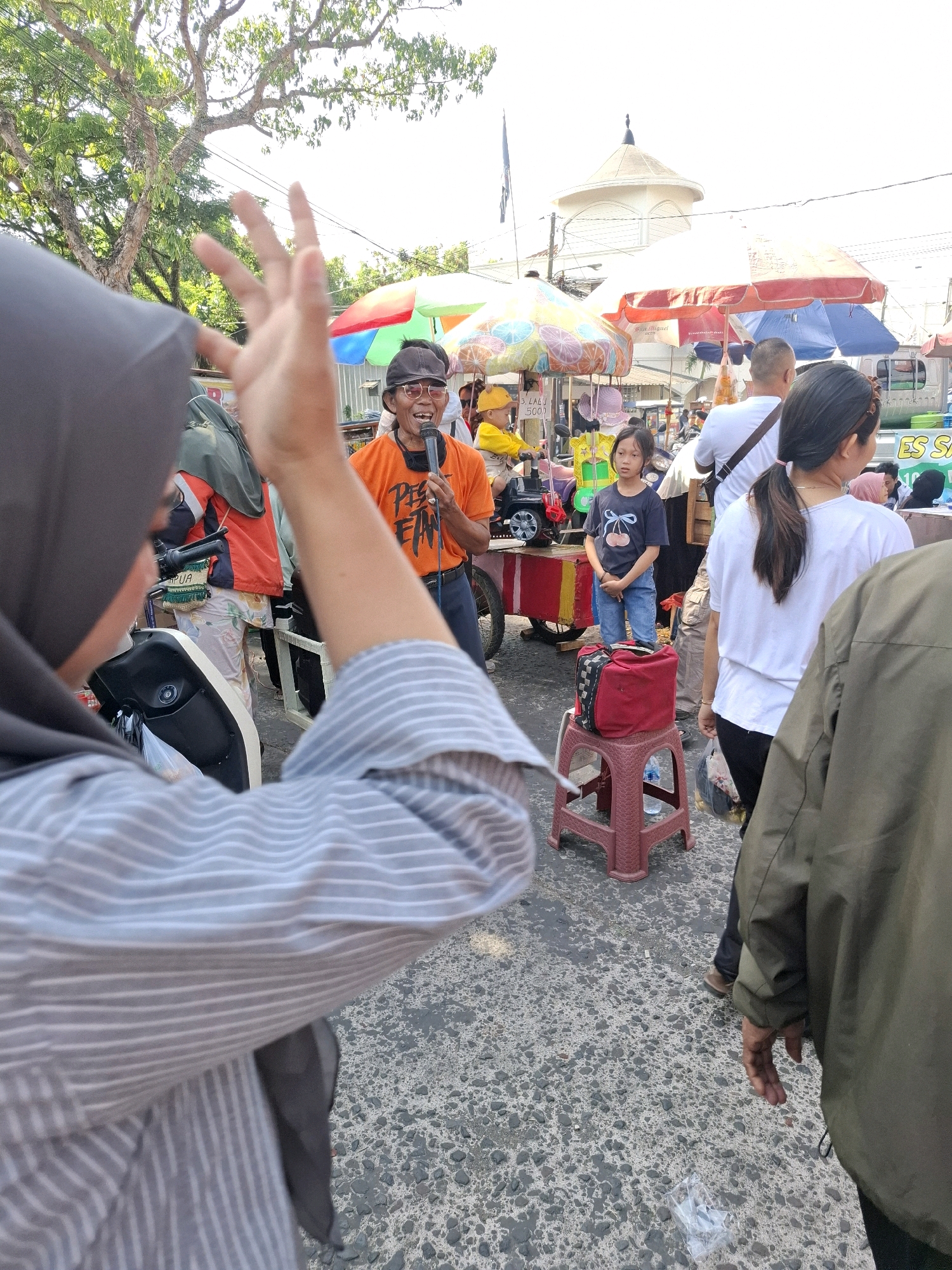 The image depicts an outdoor street scene, likely a market, in the city of Karangtengah, Indonesia. In the foreground, a person wearing a grey and white striped shirt and a dark headscarf is partially visible from the back, with their left hand raised, fingers spread. To the right of this person, a man in an orange t-shirt and sunglasses is holding a microphone and speaking, appearing to address a crowd. He has a microphone in his right hand and his mouth is open. Behind him, a young girl in a dark t-shirt with a cherry design and blue jeans stands near a red plastic stool with a red bag on it. The background shows various market stalls with colorful umbrellas, people browsing, and buildings. The ground is paved with asphalt and small stones. The overall atmosphere suggests a lively community gathering or event.