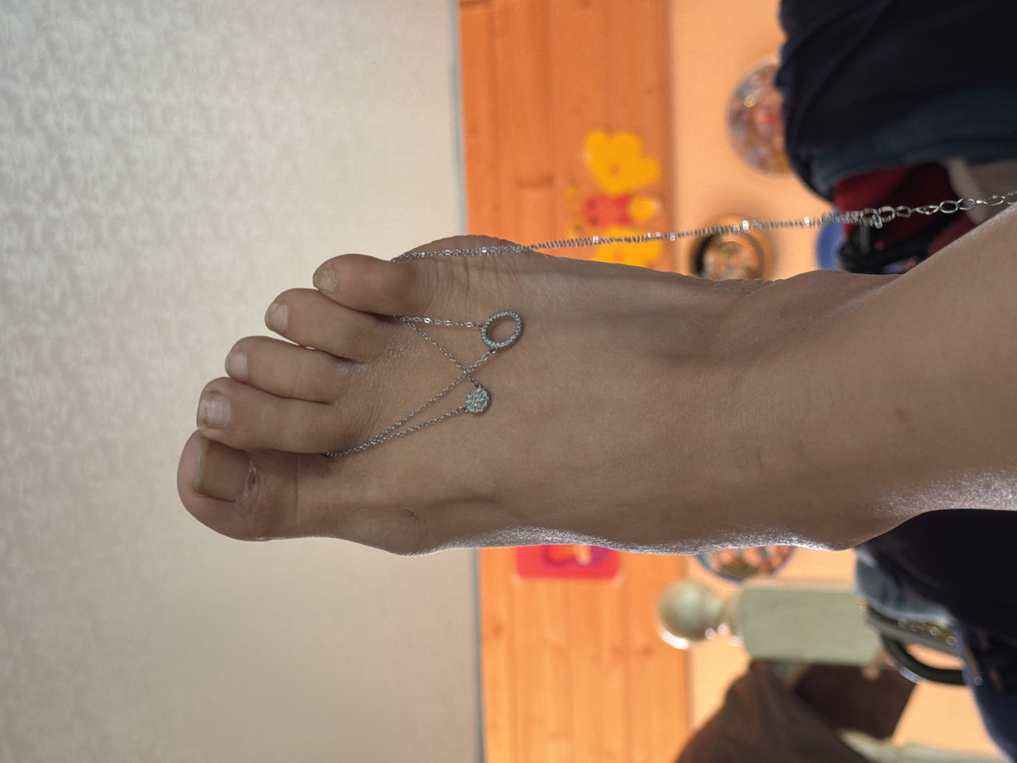 The image shows a close-up view of a person's foot, specifically the toes and the top of the foot, adorned with a delicate silver necklace. The necklace features two small pendants: one is a circular shape covered in small, sparkling stones, and the other is a thin, open circle, also adorned with similar stones. The necklace drapes across the foot and down towards the heel. The foot appears to be raised, with the toes pointing upwards. The background is blurred but suggests an indoor setting with warm-toned wooden paneling and some indistinct objects that could be furniture or decorative items. There is no identifiable information within the image that places it in Shumki, Russia.