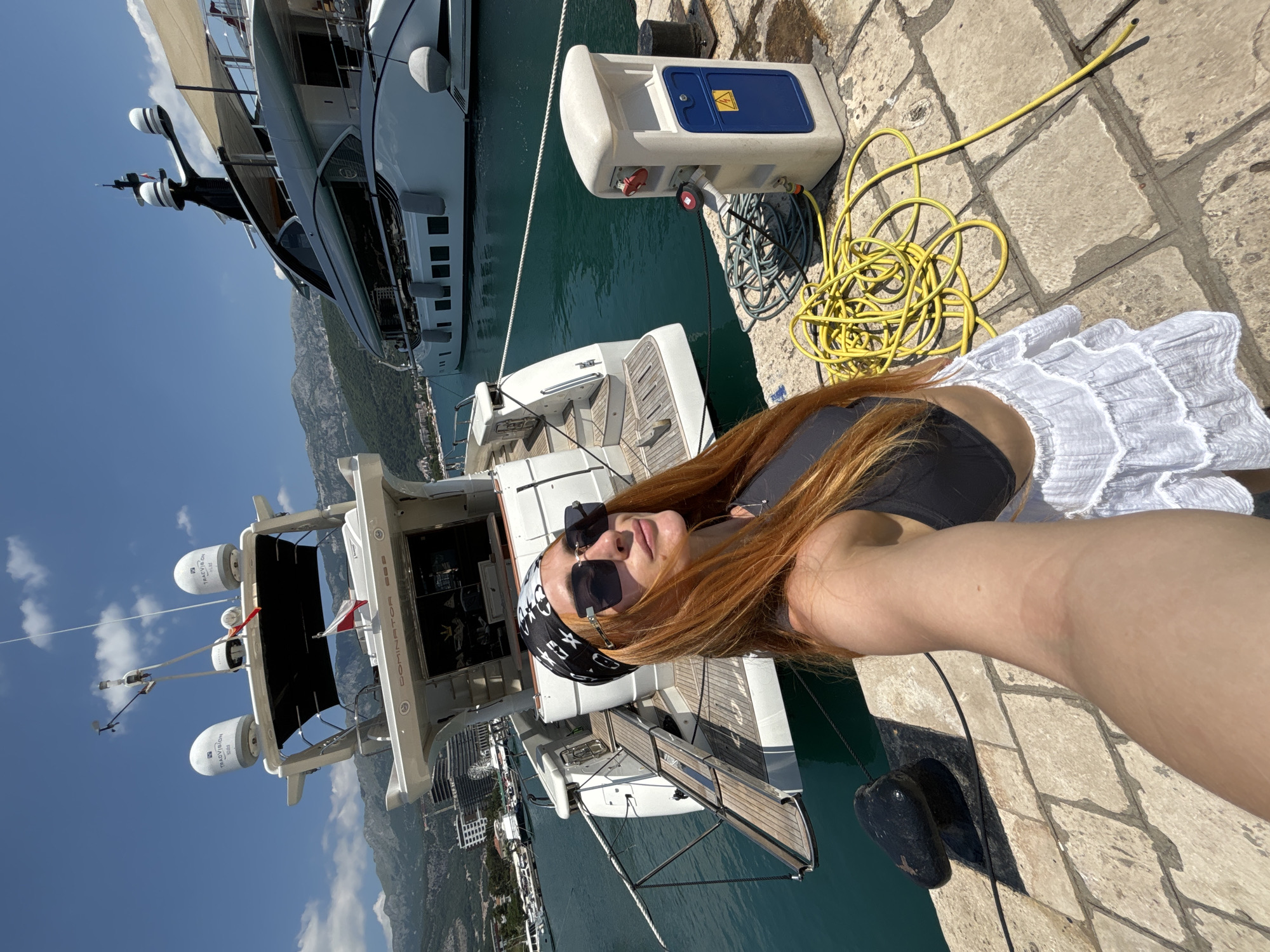 A woman with long, red hair and a bandana tied around her head is taking a selfie on a sunny day at a marina in Budva, Montenegro. She is wearing sunglasses, a dark sports bra, and a white tiered skirt. Behind her, two yachts are docked. The yacht in the foreground is smaller and white, with a wooden deck and a Polish flag. The larger yacht in the background is white with black accents and appears to be more luxurious. The marina has a stone-paved walkway, a black bollard, and a blue and white electrical pedestal with coiled yellow hoses. The water in the marina is a vibrant turquoise, and the sky is clear blue with a few scattered clouds. Mountains can be seen in the background.