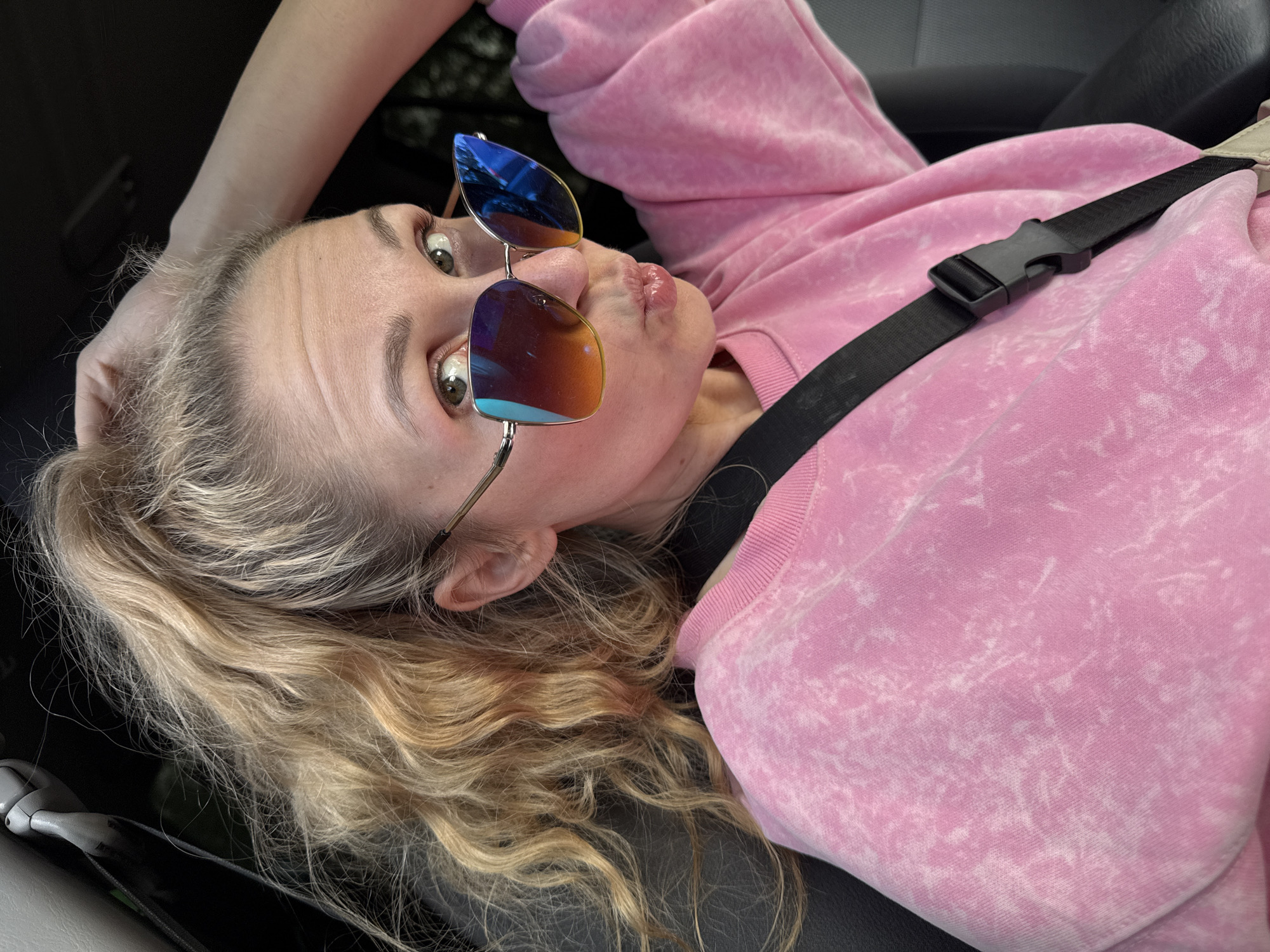 A woman with blonde hair pulled into a high ponytail, wearing a pink tie-dye sweatshirt and large sunglasses, pouts while looking at the camera. She is sitting in a car, with her hand on top of her head. A black seatbelt is visible across her chest. The background is dark and out of focus, suggesting an interior car setting. The provided crops focus on the woman's hair, face, and clothing, as well as the car's interior elements like the sun visor and seatbelt buckle. There is no information provided that can link this image to the city of Chaikovsky, Russia.