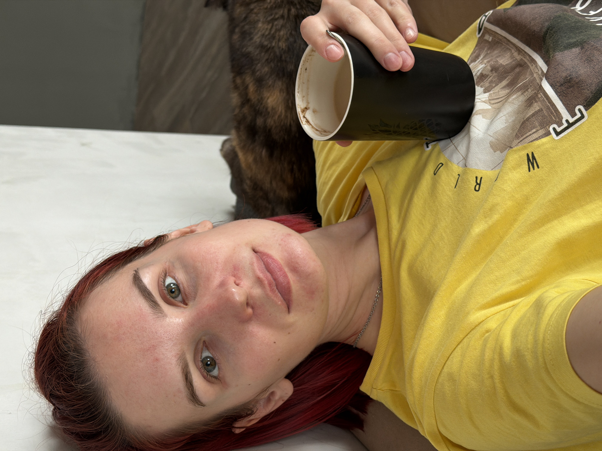 This medium-shot portrait, captured in Chaikovsky, Russia, features a person lying down and gazing directly at the camera. The image is oriented with the subject's head towards the bottom left, and her body extending diagonally upwards. She has fair skin, reddish-brown hair, and green-brown eyes. Her face displays some redness, and she has neatly groomed eyebrows and light-colored lips.
She is dressed in a bright yellow short-sleeved t-shirt with a graphic print on the chest. The print depicts a stylized globe or map in muted brown and beige tones, with visible partial letters "W", "R", "L", "D" suggesting the word "WORLD." A thin silver chain necklace is visible around her neck.
Her right hand holds a black mug with a white interior, which contains visible dried residue, likely from a finished beverage such as coffee or tea. The mug's exterior features a subtle, patterned texture. Partially visible behind the mug is a furry animal, possibly a dog or cat, characterized by its dark brown or brindle fur. The person appears to be lying on a light-colored surface in an indoor setting. The background behind the animal is an indistinct light brown or grey wall. The subject maintains a neutral expression, looking straight at the viewer.
