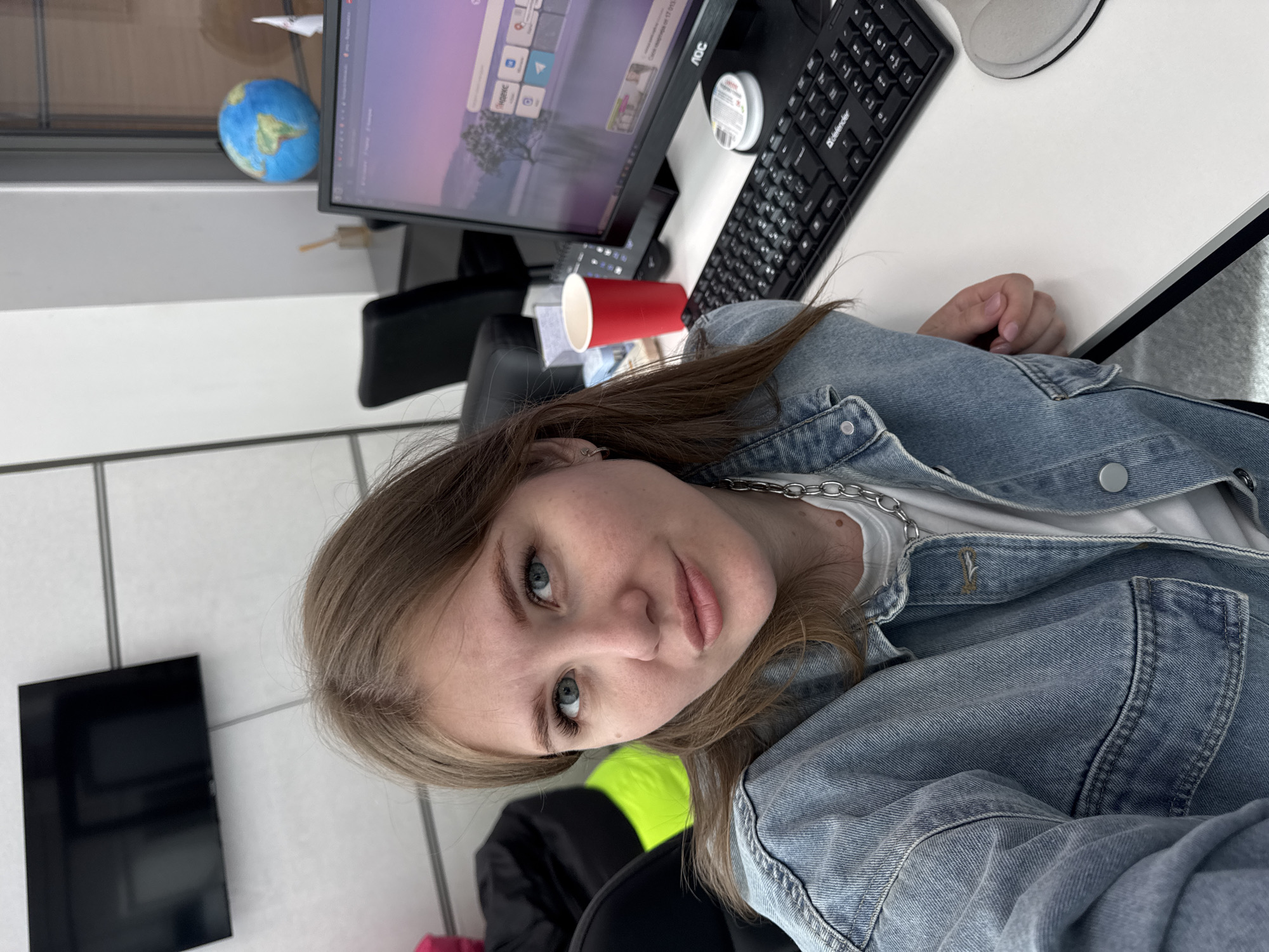 A woman with light brown hair and blue eyes, wearing a denim jacket and a silver chain necklace, sits at a desk. On the desk, there is a computer monitor, a keyboard, a mouse, and a red cup. A small globe is visible in the background near the monitor. The location is implied to be in Perm, Russia. The details suggest an office or workspace environment.