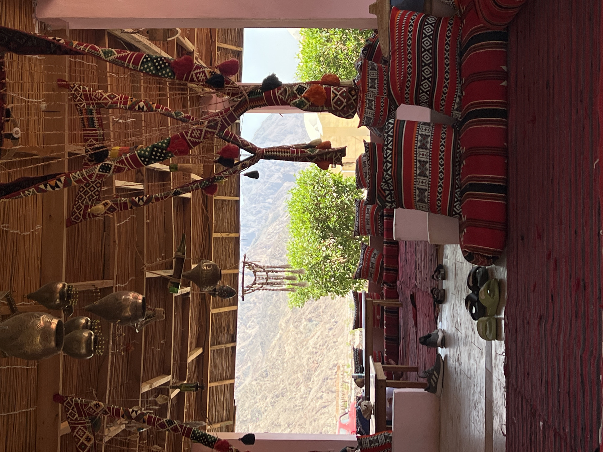 The image depicts an outdoor seating area with a traditional, rustic ambiance, likely in or near Nueiba, Egypt, given its desert mountain backdrop. The seating consists of floor cushions and low tables, adorned with red, black, and white striped fabric and geometric patterns. Several pairs of casual footwear, including sneakers and flip-flops, are scattered on the tiled floor, suggesting recent occupancy.

Overhead, a thatched roof is supported by wooden beams, from which hang various decorative elements. These include metal lanterns with an antique appearance and elaborate fabric hangings in vibrant colors and intricate patterns, some with tassels. A decorative tree-like structure, also covered in colorful, patterned fabric and tassels, stands prominently in the foreground.

Through an open-air structure, a scenic view of arid, mountainous terrain is visible, characteristic of the Sinai Peninsula. Lush green foliage partially obscures the view of the mountains, indicating a more verdant area near the seating. A glimpse of a red vehicle can be seen in the distance. The overall atmosphere is relaxed and inviting, blending indoor comfort with the natural beauty of the surrounding desert landscape.
