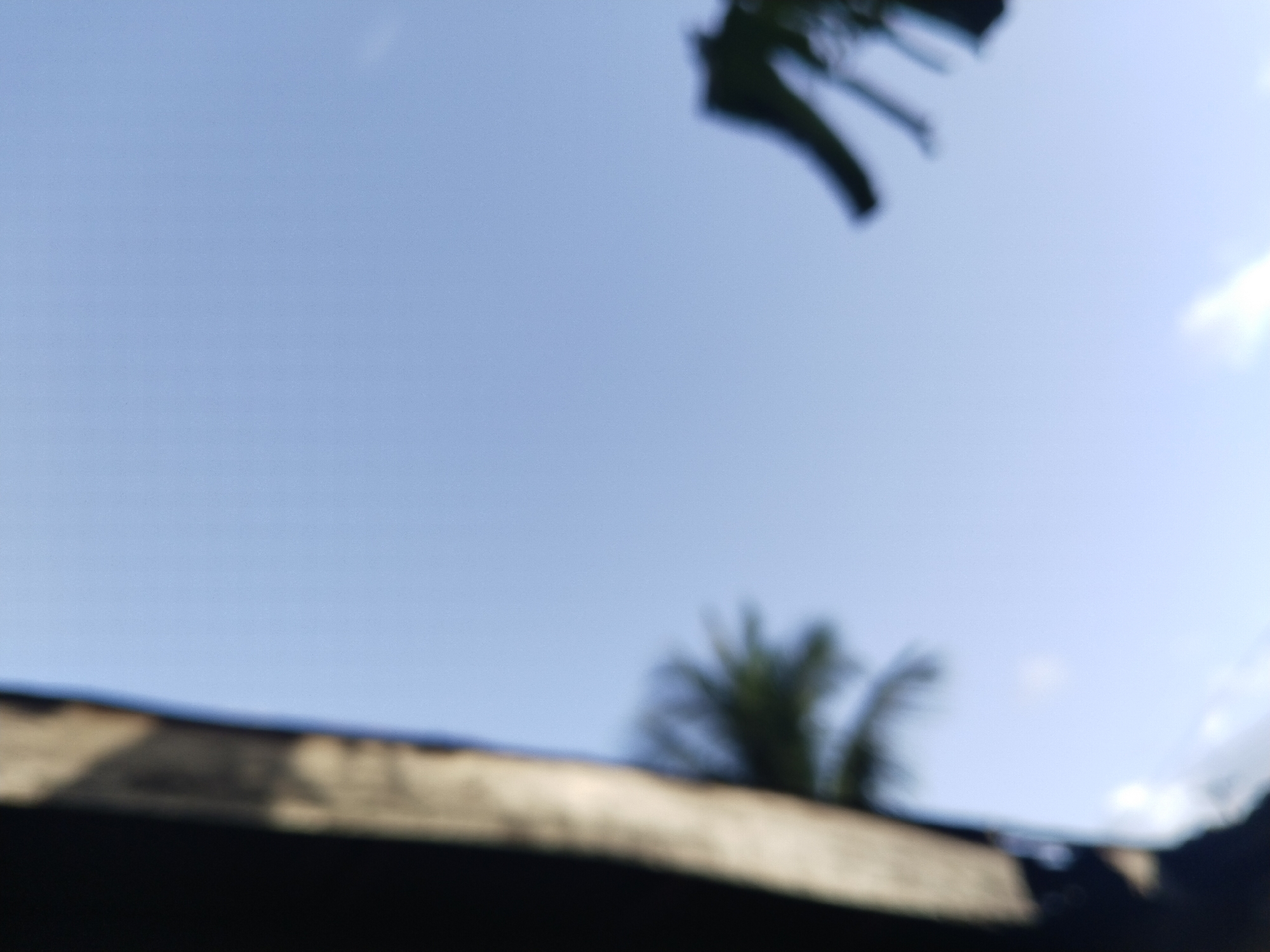 The image shows a view looking upwards, likely taken from ground level. The sky is light blue and mostly clear. A few blurry objects are visible in the foreground and lower portions of the frame. These seem to include parts of a roof or structure, as well as foliage from a palm tree and other plants. Given the prompt and context, it's probable the location is within Uyo, Nigeria, but specific details for exact localization are insufficient.