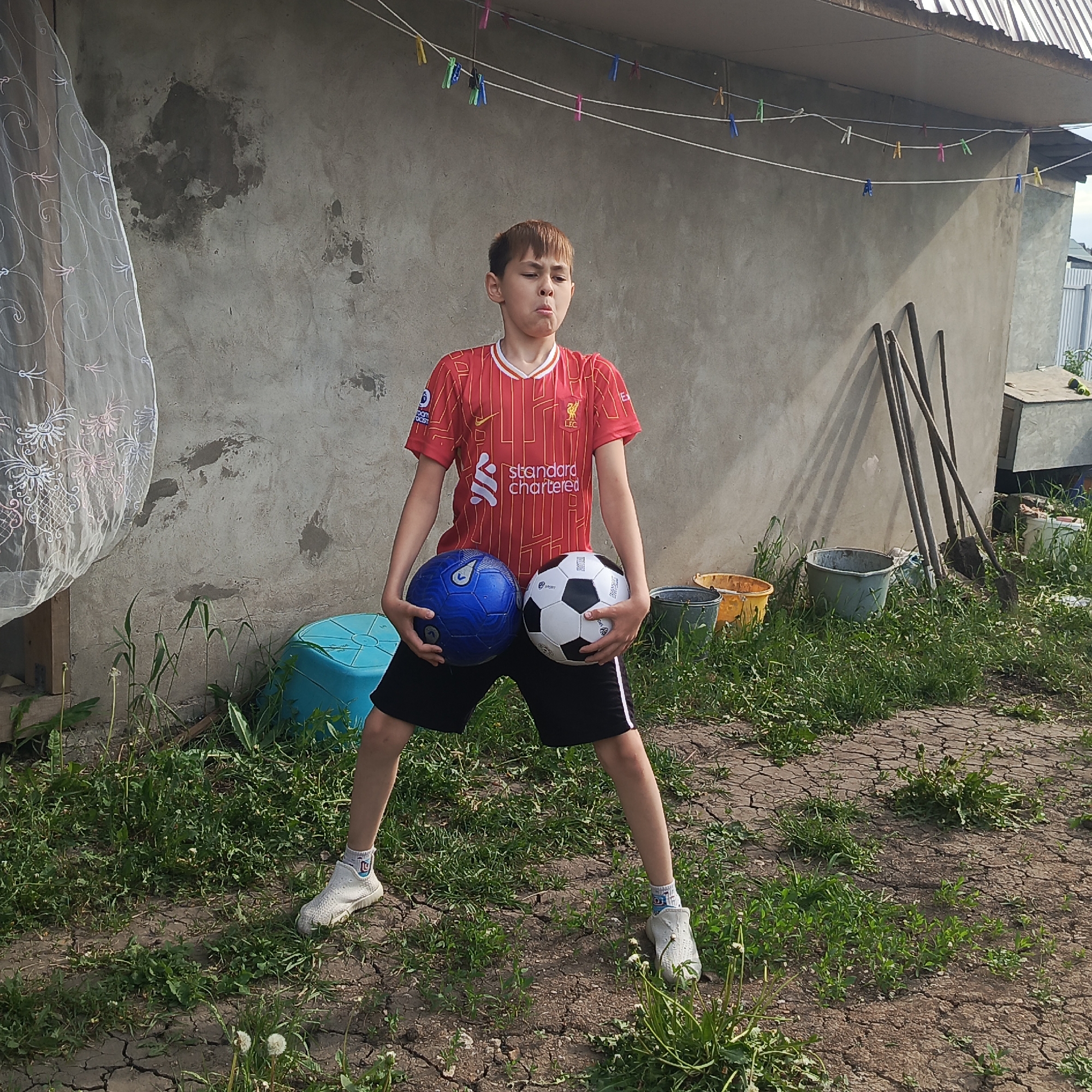 A young boy stands in a yard in an unknown location, holding a blue soccer ball in his left arm and a black and white soccer ball in his right. He wears a red Liverpool jersey and black shorts with a white stripe. His mouth is puckered, and his gaze is directed upwards. He has short brown hair and is wearing white athletic shoes. The yard is a mix of cracked dirt and grass. Behind him is a concrete building with a clothesline strung with clothespins. Various buckets and garden tools are visible in the background, adding to the cluttered outdoor scene. A white curtain hangs to the left side of the frame.