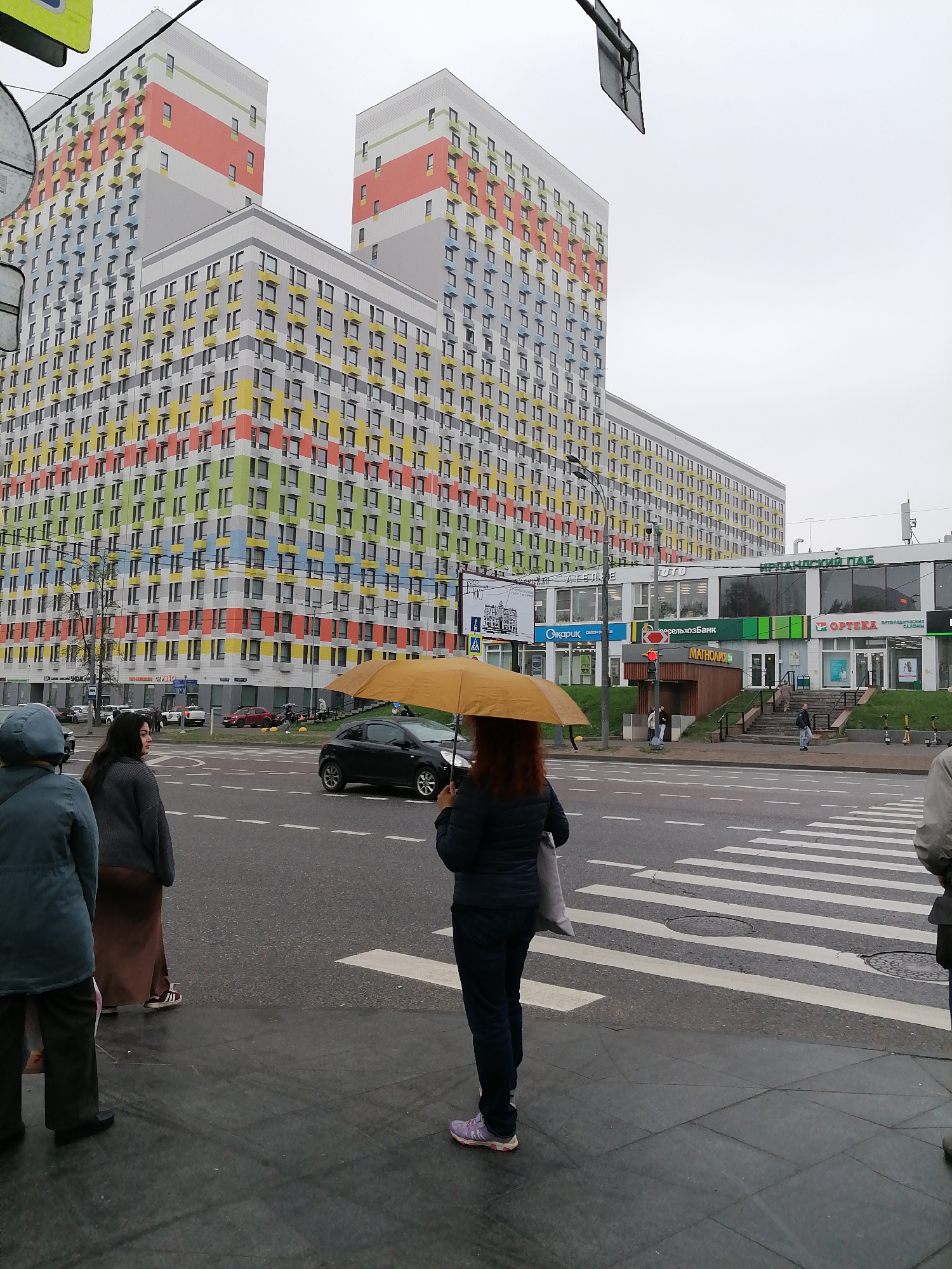 The image shows a street scene in Moscow, Russia, characterized by modern, high-rise apartment buildings with colorful facades in the background. In the foreground, several pedestrians are standing on the sidewalk and at a crosswalk, appearing to wait. One person is holding an open yellow umbrella. A black car is visible on the street, crossing the pedestrian walkway. Across the street, there is a commercial building with several retail stores and businesses. The weather appears overcast and possibly rainy.