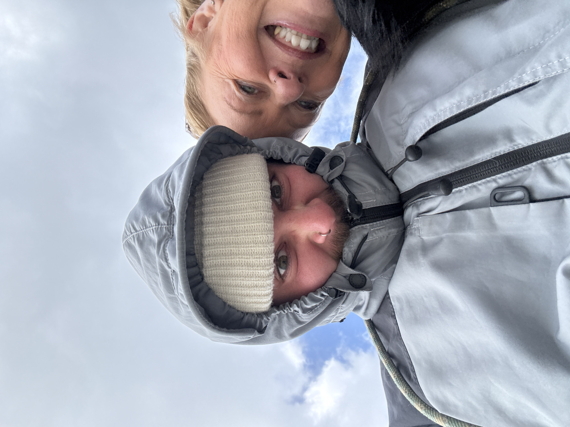 Two individuals are closely positioned in the image. The individual on top has blonde hair, fair skin, and is smiling broadly, showcasing their teeth. They have a nose ring and visible wrinkles around their eyes. The person below is wearing a light grey hooded jacket with the hood up, revealing a white knitted beanie underneath. Their face is partially obscured by the hood and beanie. The background consists of a cloudy sky. The location and city are not discernible from the image.