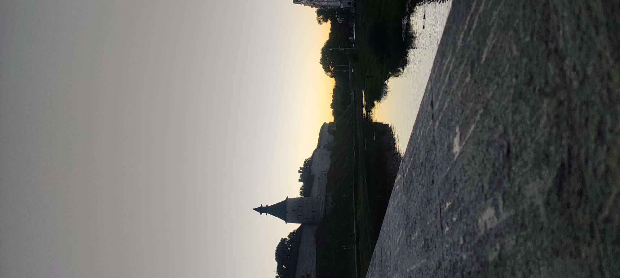 This is a vertical, medium-distance, eye-level shot of the Pskov Kremlin in Pskov, Russia, at dusk. The sky is a gradient of pale gray at the top to a soft yellow-orange at the horizon. In the mid-ground, a large, stone fortress wall runs horizontally across the frame, with a prominent, conical-roofed tower on the left. The fortress is partially silhouetted against the sunset. A dark river flows in front of the fortress, reflecting the sky and the silhouette of the wall. The foreground is dominated by a textured, gray stone surface, possibly a bridge or embankment, angled diagonally from the bottom left to the top right. Trees are visible along the riverbank and on the fortress grounds. Two ducks are swimming in the river. The overall atmosphere is serene and somewhat melancholic due to the low light and muted colors.