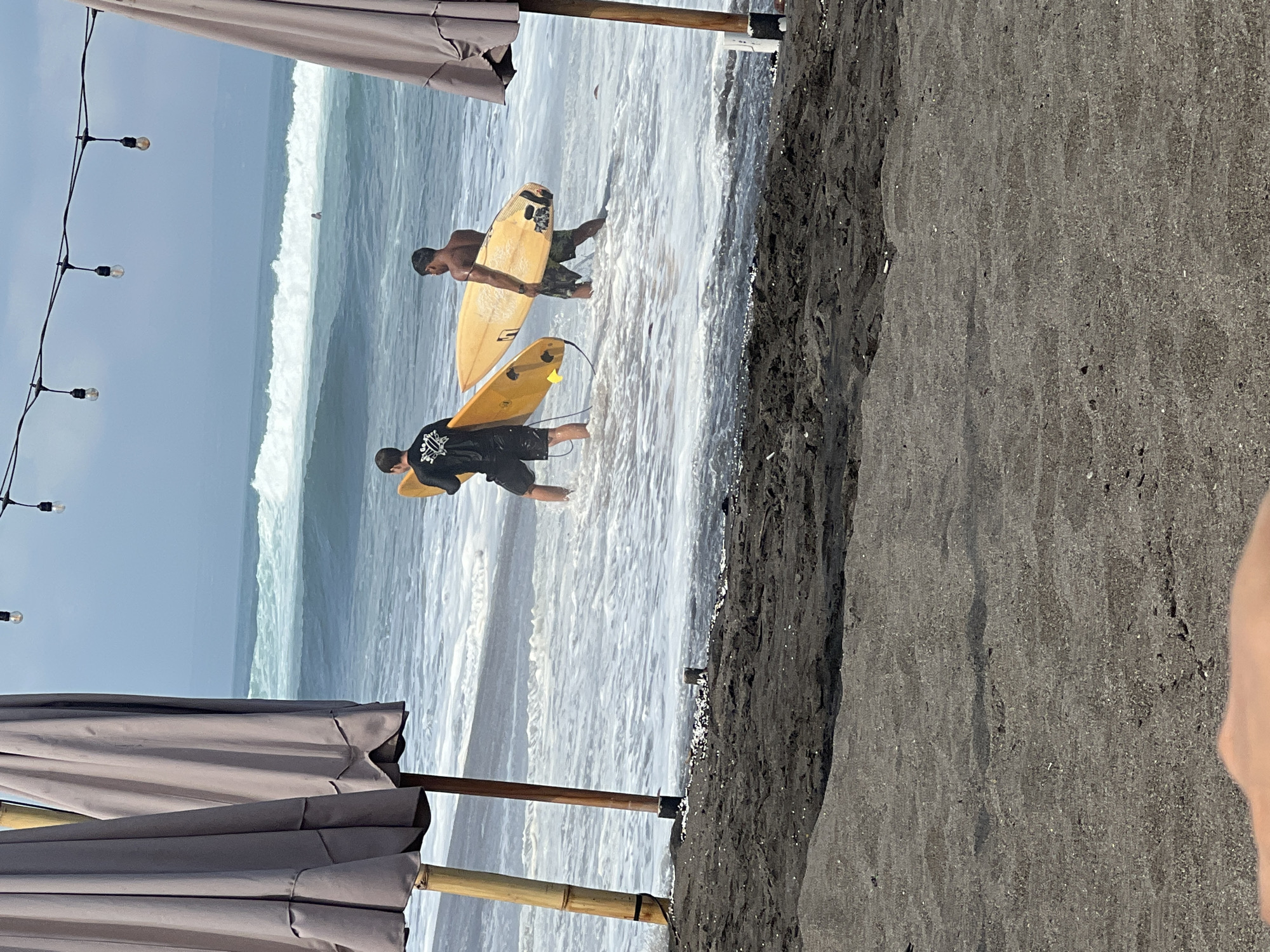 Two men walk into the ocean carrying surfboards. The man on the left wears a black shirt and shorts, and the man on the right wears patterned shorts. The ocean has large waves and foam, suggesting a surfable condition. The beach is dark, volcanic sand. In the foreground, two large beach umbrellas and bamboo poles suggest a beachside establishment. Overhead, a string of bare lightbulbs hangs, possibly for evening ambiance. In the distance, a lone figure can be seen in the ocean, further out. The overall scene depicts a beach on a sunny day, likely in a surf destination. This scene is in North Kuta, Indonesia, a region known for its surf beaches.
