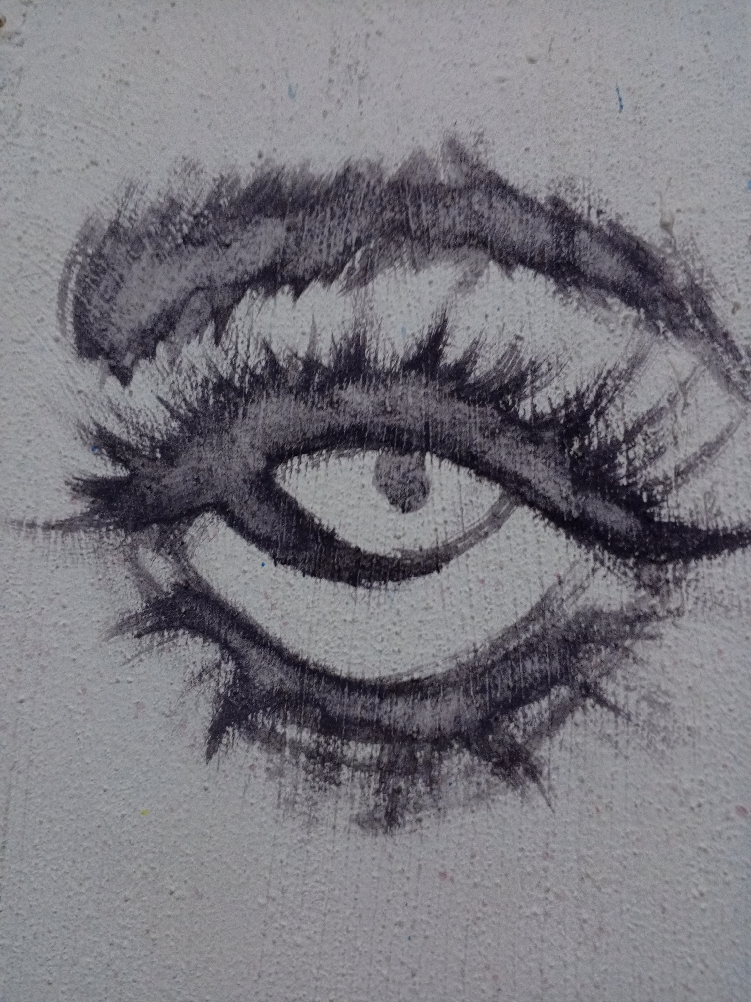 The image is a close-up of an eye painted in charcoal on a textured, likely concrete, wall. The eye is stylized with thick, expressive eyebrows and eyelashes, and a visible iris. The artwork appears to be part of a larger mural or graffiti piece, given the texture of the wall, but only the eye is visible within the frame. The location, Parroquia Ocumare, Venezuela, is inferred, and there are no observable people or interactions in the scene.