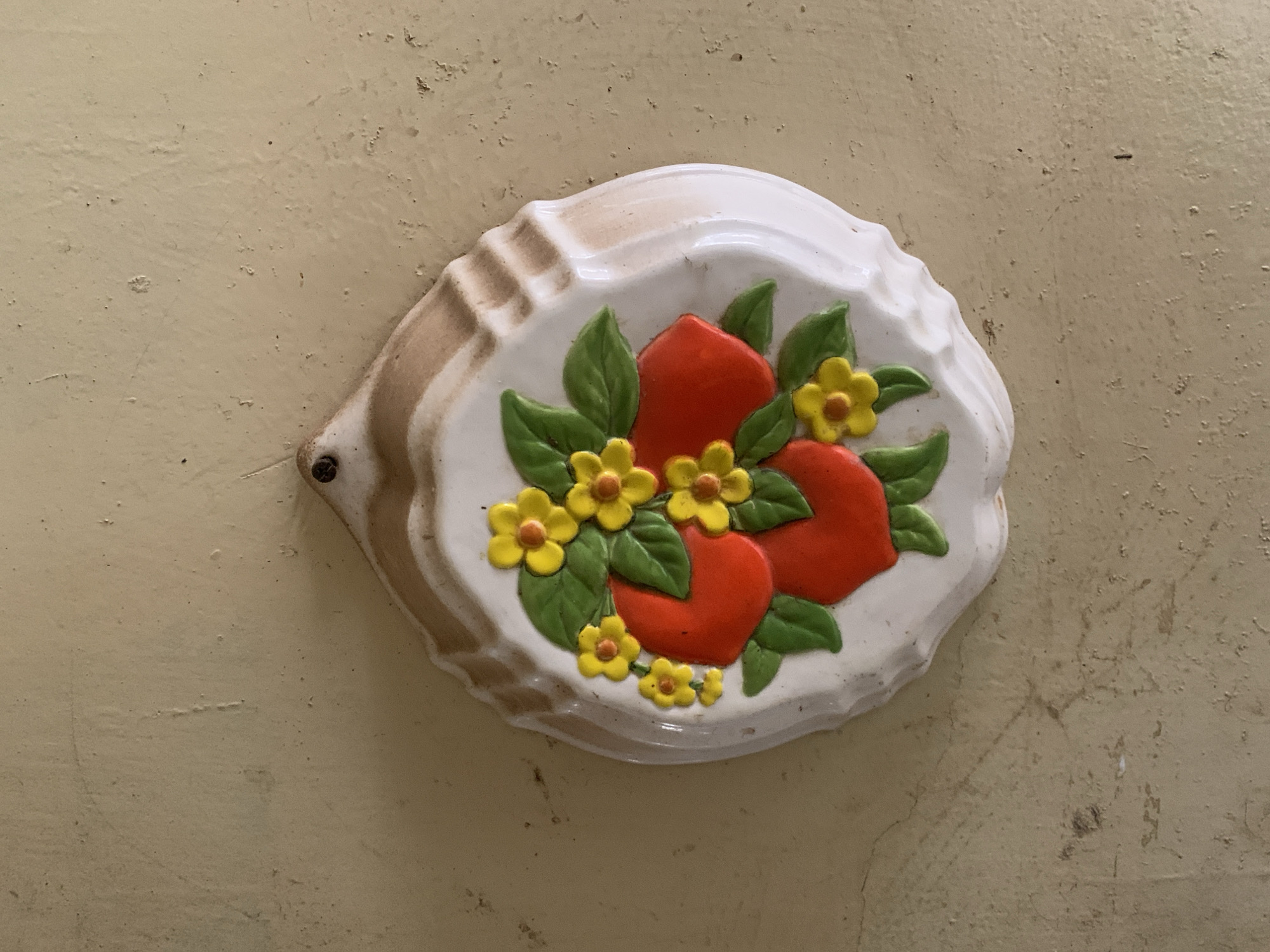 A decorative, off-white ceramic plaque is centrally featured, mounted against a worn, yellowish wall. The plaque has a wavy, scalloped edge with subtle brown accents, suggesting a vintage or rustic aesthetic. On its surface, three stylized red fruits, possibly peaches or apples, are depicted with green leaves and clusters of small, five-petaled yellow flowers with orange centers. The fruits and foliage are raised, giving the plaque a three-dimensional quality.
A dark screw or nail is visible in the upper left corner of the plaque, securing it to the wall. The wall itself exhibits signs of age and wear, with varying textures, small indentations, and potential areas of flaked paint or accumulated dust, contributing to a lived-in feel. The lighting is even, highlighting the details and colors of the ceramic piece. There is no visible text or specific activity occurring in the image, which captures a static decorative object in a domestic or semi-public setting. The overall scene suggests an indoor environment.