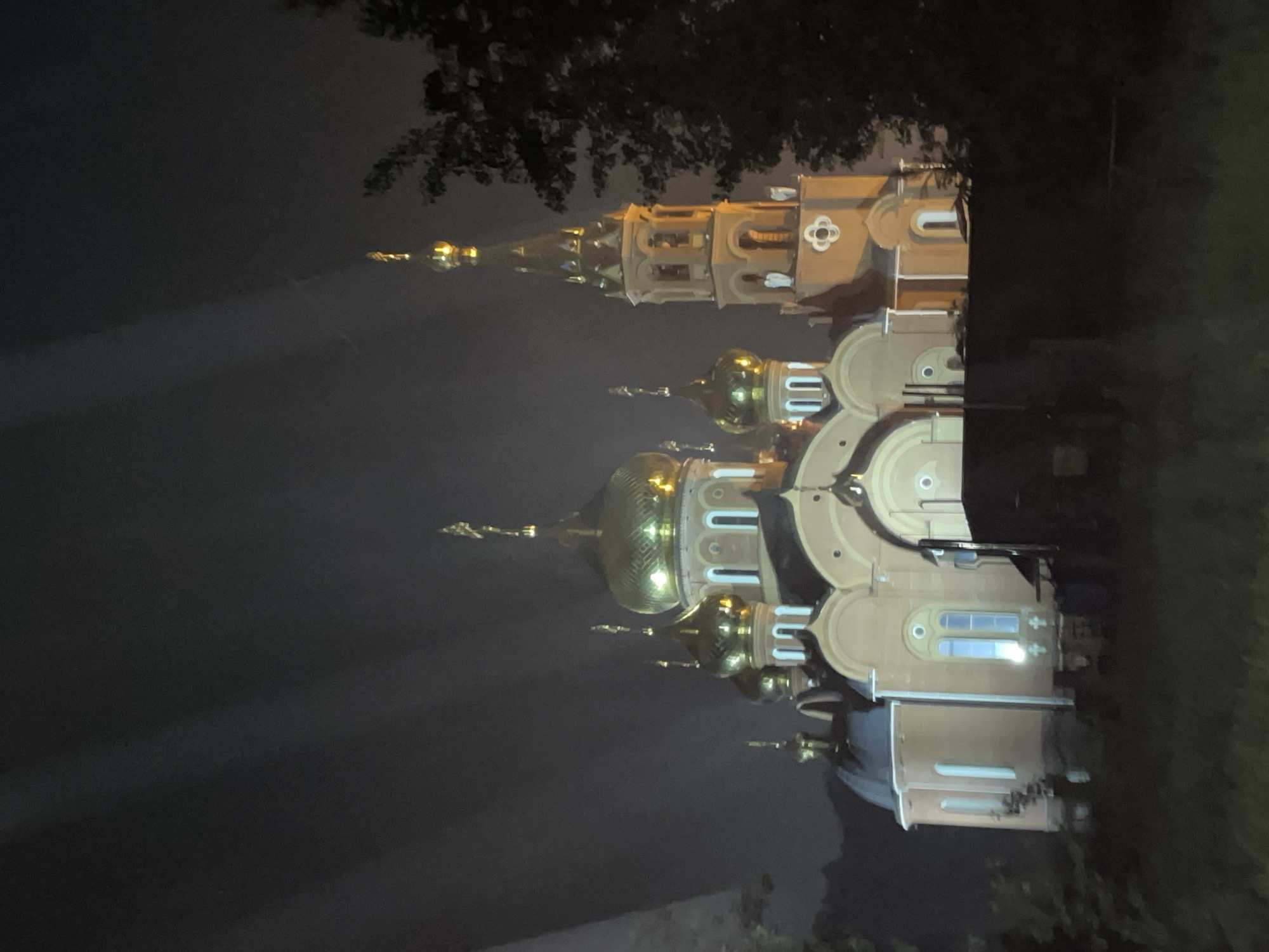 The image displays the Church of St. George the Victorious in Chaikovsky, Russia, captured at night. The Russian Orthodox church features a light beige or peach-colored facade with distinct white architectural trim around its numerous arched windows and structural elements. Five prominent golden onion domes, each topped with a Russian Orthodox cross, are visible on the main structure, with a larger central dome flanked by four smaller ones. To the right, a tall, multi-tiered bell tower with a golden conical spire and cross rises, featuring arched openings for bells. The entire edifice is brightly illuminated by external and internal lights, casting a warm glow. The sky above is dark and cloudy, with faint, parallel light streaks visible in the upper left portion of the frame. Dark, indistinct tree foliage partially obstructs the view on the far right and in the unlit foreground, which appears to be ground covered in low vegetation. No individuals or specific actions are discernible.
