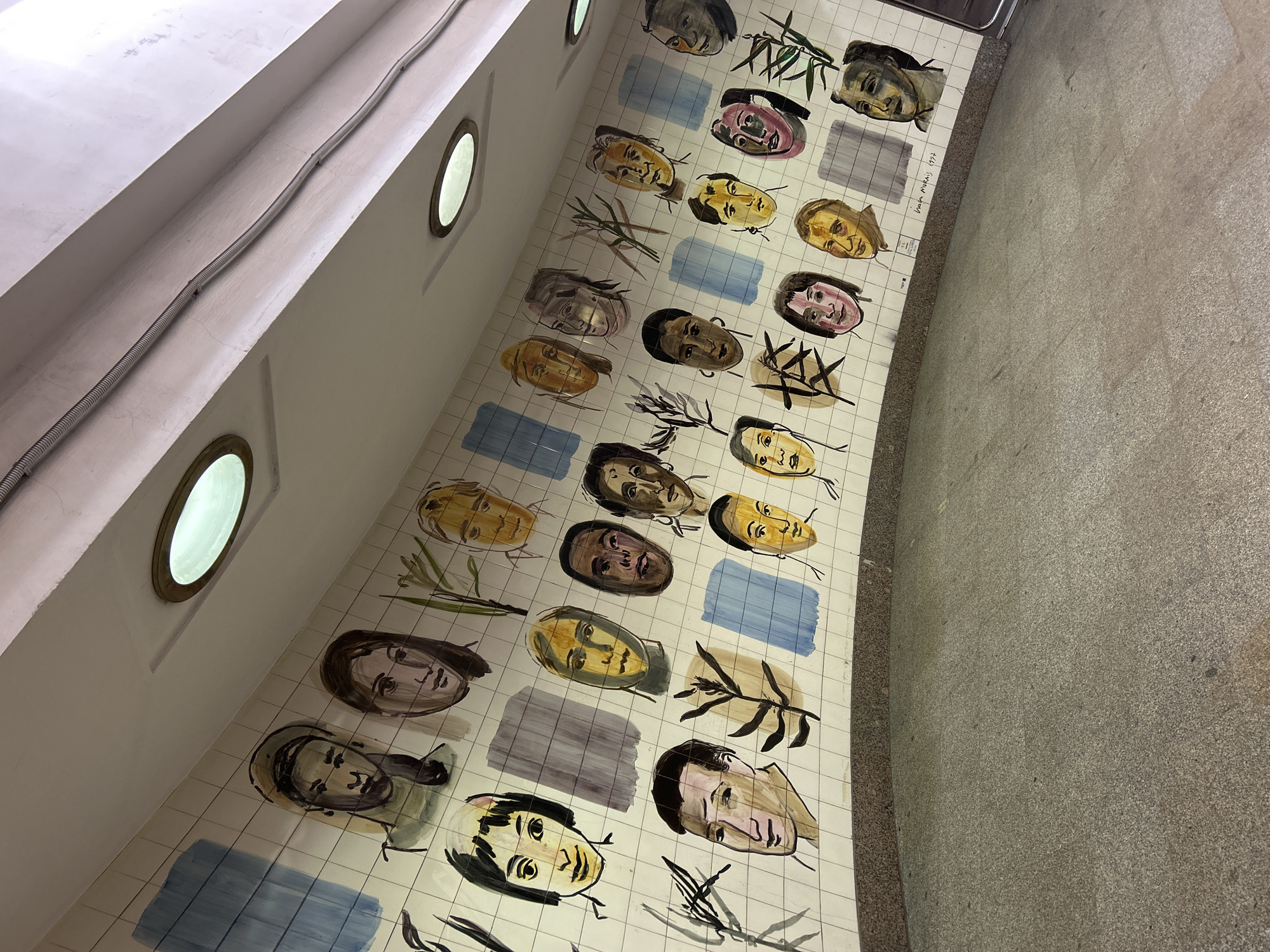 The image shows a curved wall tiled with painted faces and blue, gray, and white tiles. Small porthole windows are present on the wall above the tiled section. The tiled wall curves to meet a gray concrete floor. A signature and the date "1992" are visible next to one of the faces, indicating it is likely an artwork. The location is Unknown.