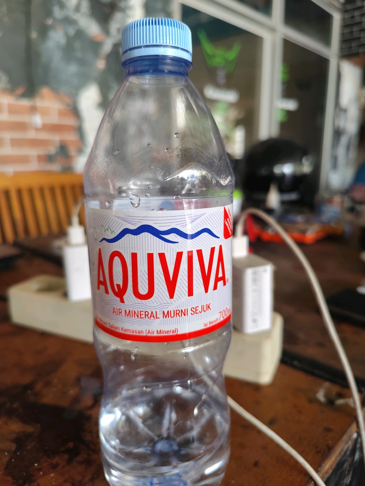 A clear plastic bottle of Aquviva mineral water is positioned centrally on a wooden table. The bottle has a blue cap and a white label featuring red text that reads "AQUVIVA AIR MINERAL MURNI SEJUK" and "Isi Bersih 700ml". Condensation is visible on the outside of the bottle. In the background, a power strip with three white electrical adapters is partially visible. The environment appears to be indoors, possibly a cafe or an office space, with blurred elements suggesting furniture and a window in the distance. There are no people or animals present. The image quality is good, with sharp focus on the bottle. The scene is typical of an everyday setting, likely in Palangka Raya, Indonesia, given the brand name and typical Indonesian settings.