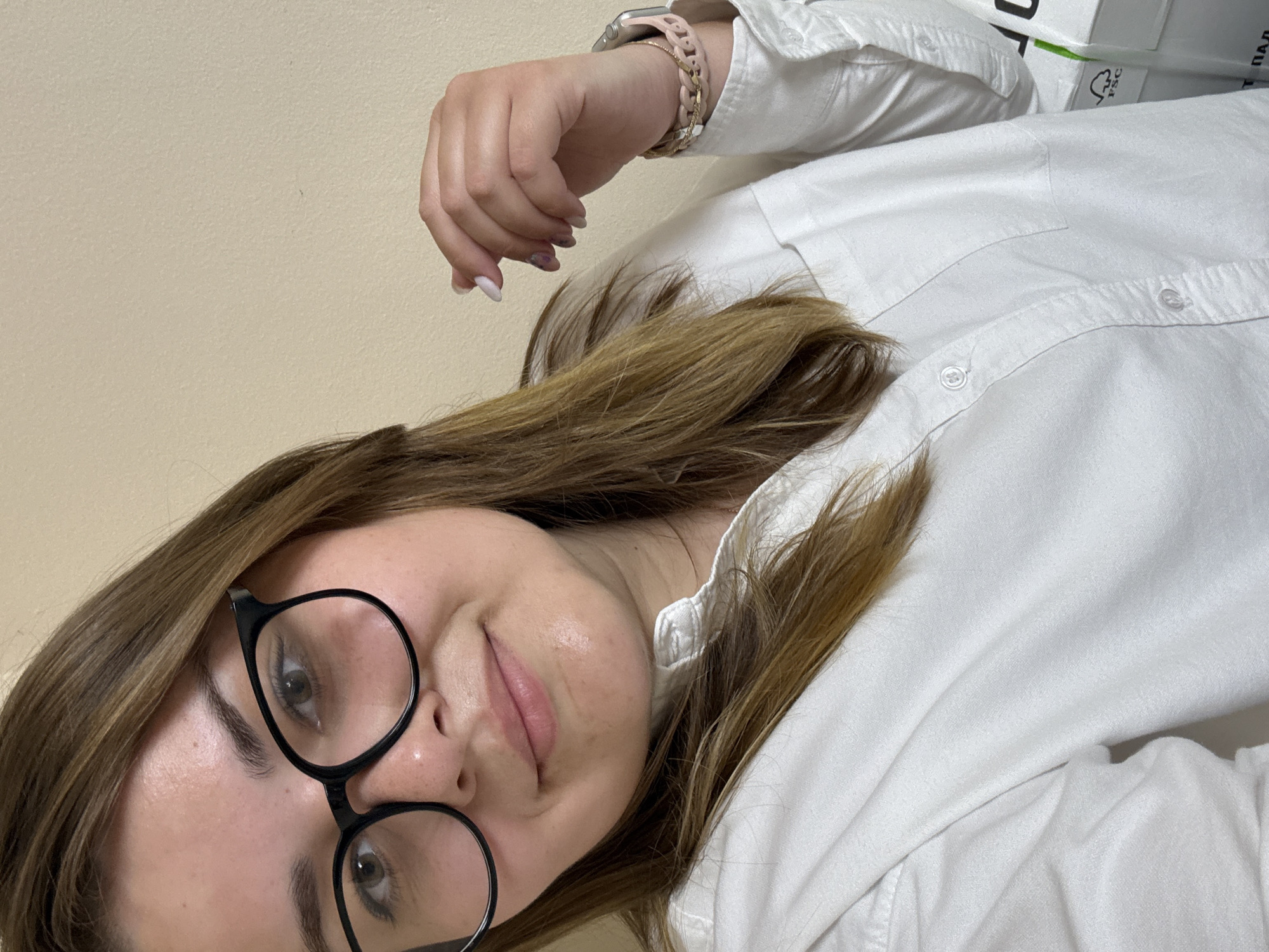 A woman with light brown hair and black-rimmed glasses is facing the camera. She is wearing a white button-down shirt. She has a watch on her left wrist. The background is a plain, light-colored wall. There is a stack of white paper in the background. The image location cannot be determined.