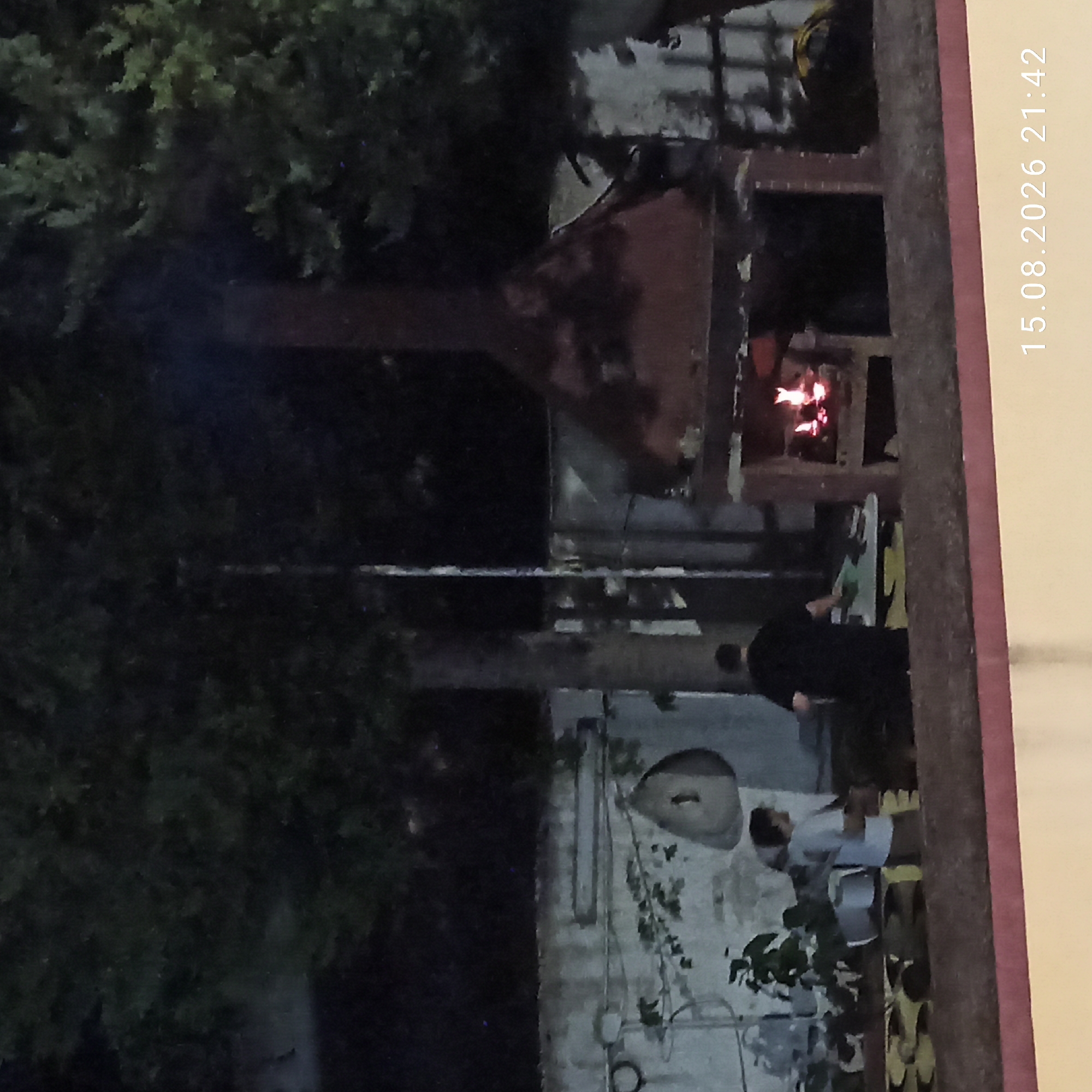 This is a night-time, eye-level, medium shot photograph taken outdoors. In the center of the image is a large, brick barbecue with a fire burning inside. To the left of the barbecue, two individuals are visible. One, wearing a light-colored shirt and dark pants, is facing away from the camera and appears to be interacting with a smaller individual. The smaller individual, wearing a white t-shirt and dark shorts, is also facing away from the camera and seems to be looking towards the barbecue. Behind them, a white wall is visible, decorated with what appears to be plants and possibly some circular objects. The background is dominated by dark trees and foliage. The overall setting suggests a backyard or patio area. The bottom right corner of the image displays a timestamp: "15.08.2026 21:42". The location is the city of Bitonto, Italy.