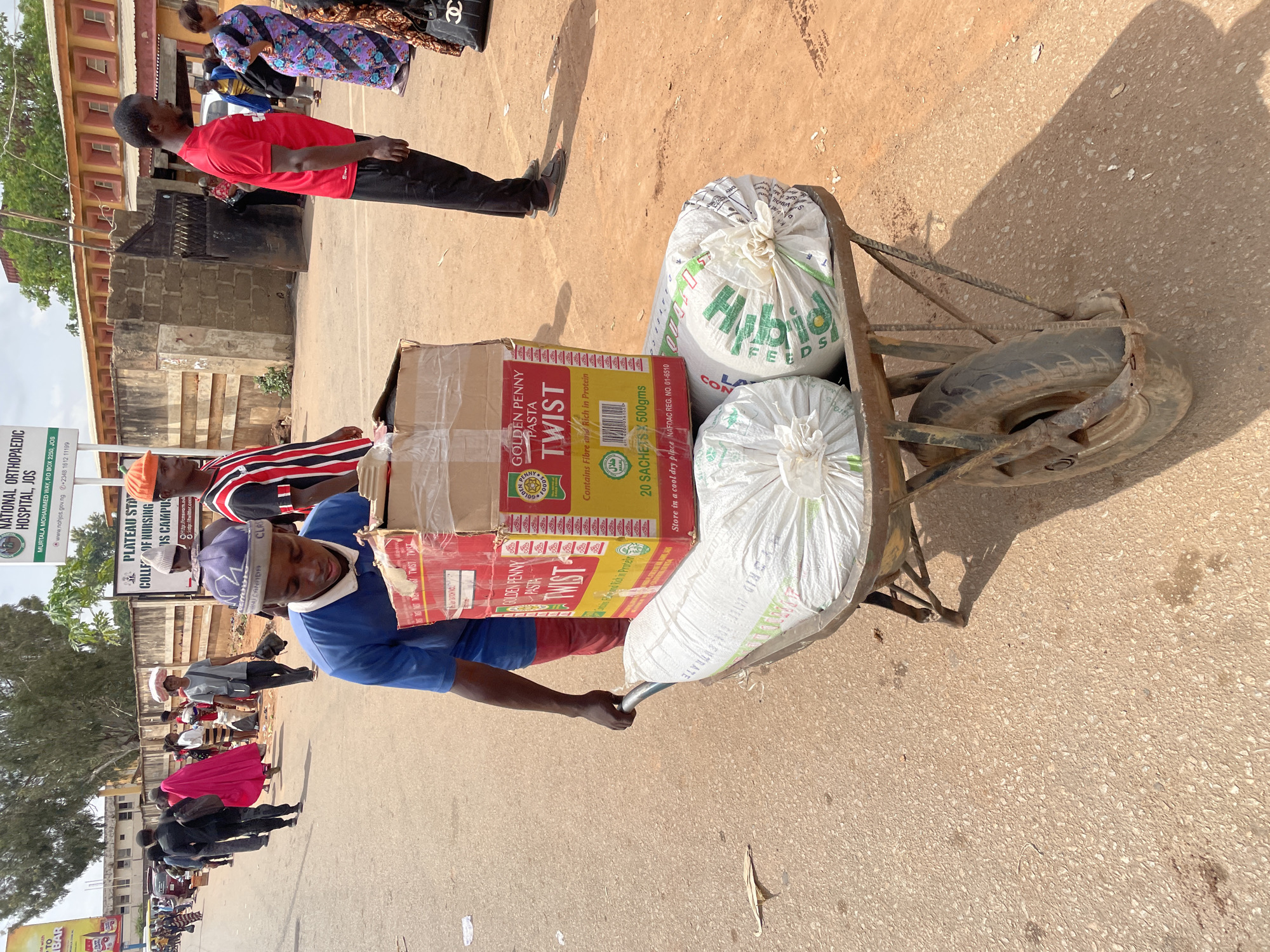 The image shows a man pulling a wheelbarrow laden with goods on a street in Jos, Nigeria. The goods include sacks labeled "Hybrid Feeds" and boxes of "Golden Penny Twist" pasta. The wheelbarrow is old and rusted. The man is wearing a blue shirt and a purple hat. In the background, there are other people walking on the street, and signs for "National Orthopaedic Hospital, Jos" and "Plateau State College of Nursing, Jos Campus" are visible. The environment appears to be a busy street with buildings and trees in the background.
