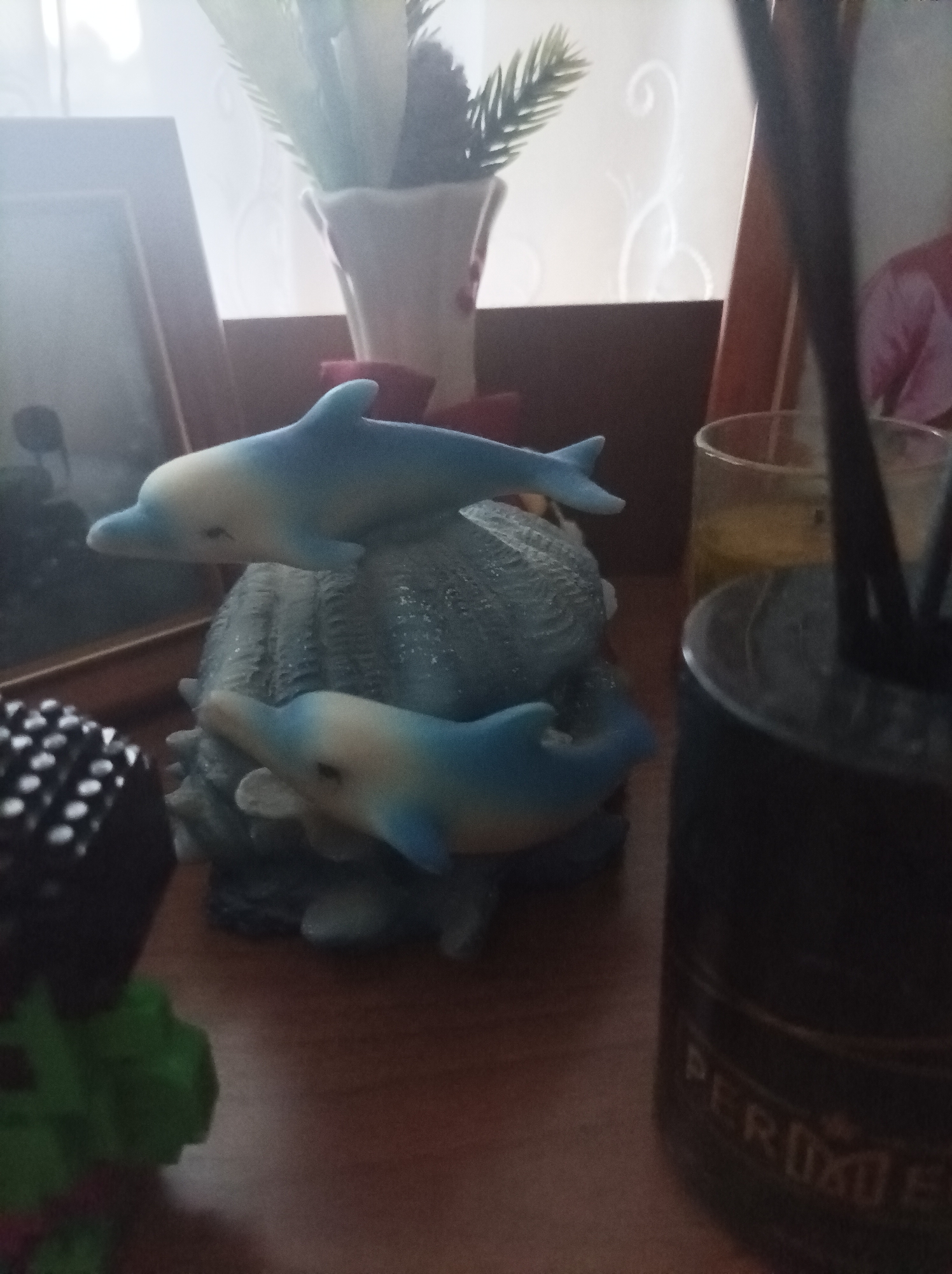 This image presents a still life arrangement on a wooden surface, likely a table or shelf, in an indoor setting in Semyonov, Russia. The central subjects are two decorative dolphin figurines, primarily light blue with creamy white undersides, resting on a textured, greyish-blue base resembling a seashell or coral. One dolphin is positioned higher, as if leaping over the other.
In the background, bright, diffused light streams from what appears to be a window, partially obscured by a white conical vase holding artificial green foliage. A wooden picture frame is visible on the left, reflecting the ambient light. On the right, another wooden frame is partially seen alongside a glass containing a yellowish liquid. Next to the glass is a dark, cylindrical container, likely a reed diffuser, with several dark sticks protruding. Faint text on this container shows partially visible letters that could spell "PEROXU" or "PERFUMES."
In the foreground on the left, a black object adorned with a grid of white circular dots, resembling a decorative cube or a die, is partially visible, along with a blurry green object beneath it. The scene is brightly illuminated from the background, suggesting it is daytime. There are no people or specific activities taking place, and no explicit emotional cues are conveyed. The overall atmosphere is tranquil.