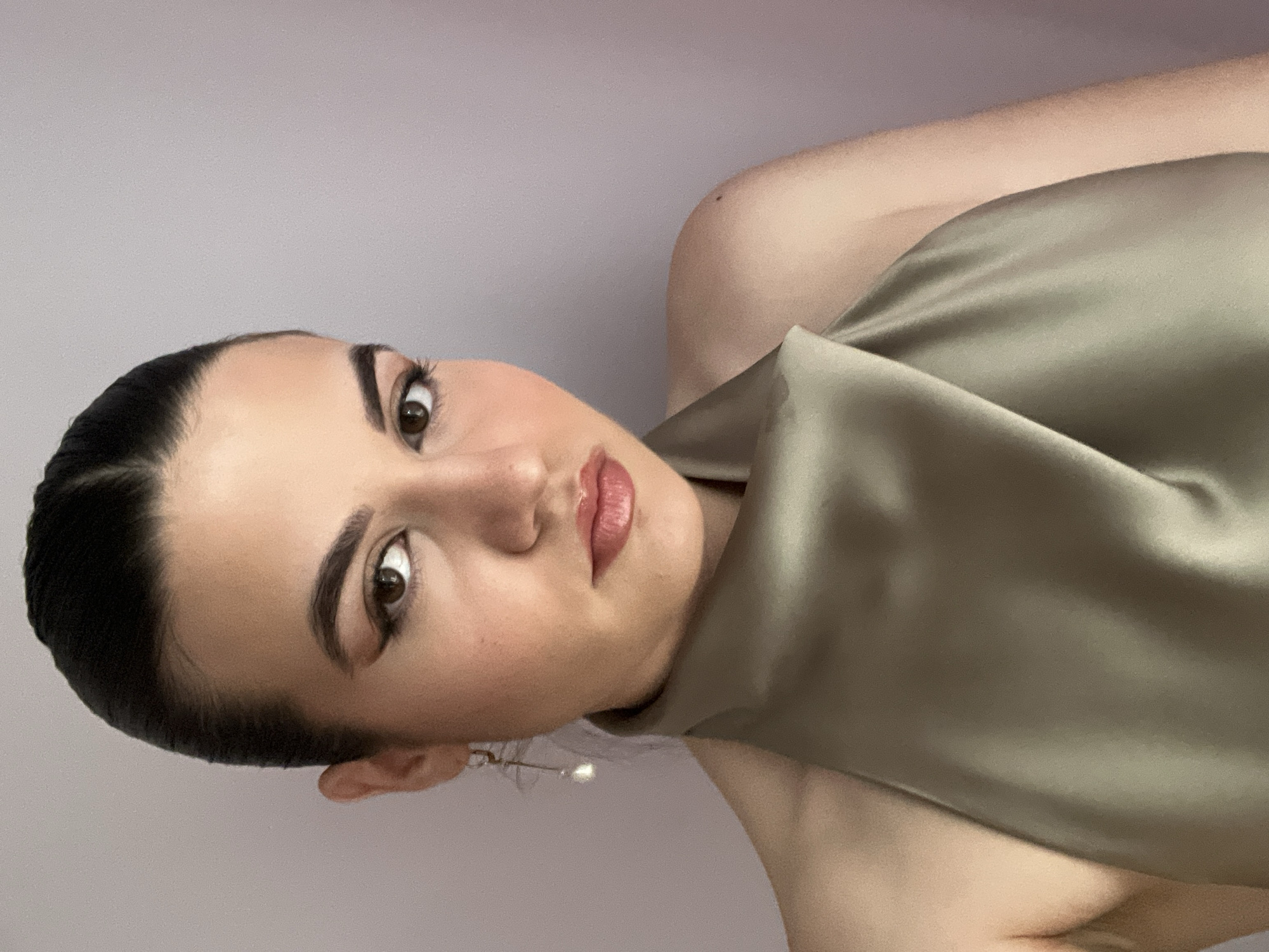 A woman with dark hair pulled back poses against a solid light purple backdrop. She wears a shiny, draped garment in a muted olive-green color. Her makeup is polished, with defined eyebrows, eyeliner, and a glossy lip. A single pearl earring is visible. The location is not specified.