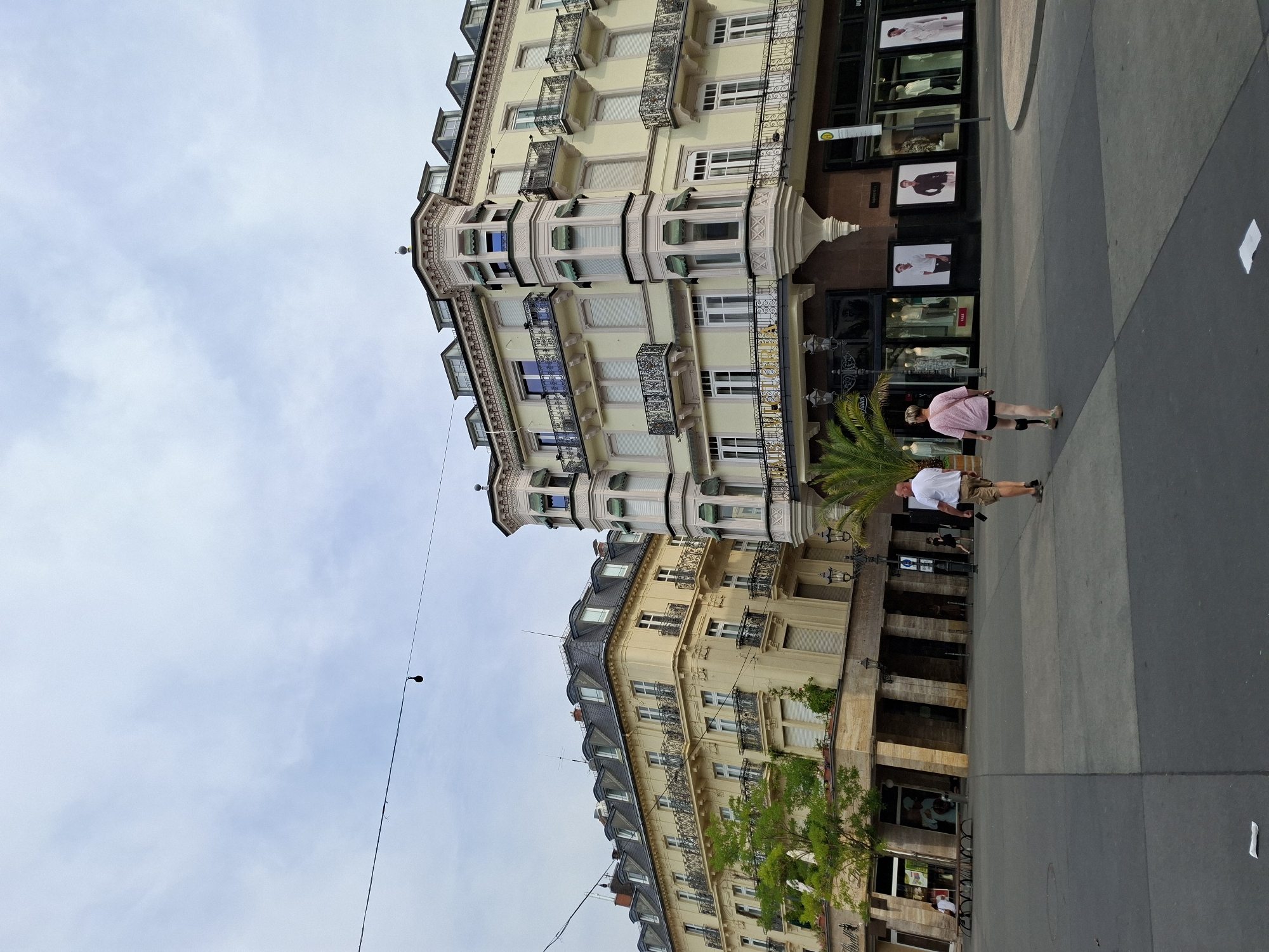 The image depicts an outdoor urban scene with two large, ornate buildings on either side of a wide, paved walkway. The building on the left is pale yellow with a mansard roof and numerous windows with decorative wrought-iron balconies. The building on the right is also pale yellow and features bay windows with green awnings and similar balconies. Storefronts are visible at the base of the right building, displaying mannequins in retail windows. A palm tree stands in front of the right building. Two people are walking away from the camera on the walkway. A man in a white t-shirt and khaki shorts is on the right, and a woman in a pink shirt and black shorts is to his left. The sky is overcast.