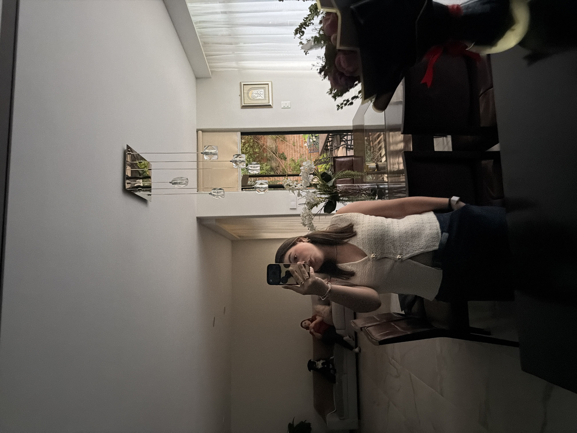A young woman is taking a selfie in a mirror. The woman is wearing a white shirt and dark pants. The mirror is in a room with a dining table. There is a bouquet of flowers on the table. The room is in a building in Barrio de Nuevo León, Mexico. There is a modern style pendant light on the wall. There is a picture hanging on the wall above the doorway. A small dog is resting on a chair in the background.