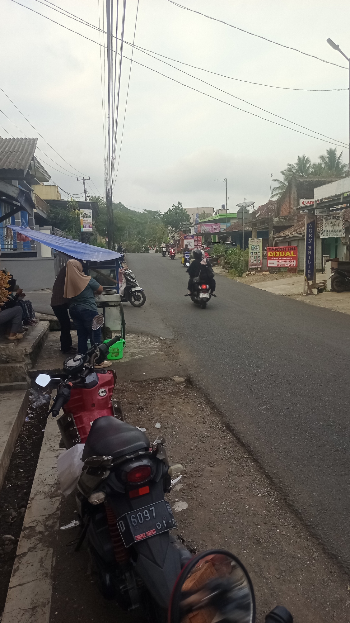 A cloudy day scene depicts a street in Karyamandala, Indonesia. In the foreground, two motorcycles are parked. A red motorcycle is on the left, and a black motorcycle with license plate D 6097 is in the center. Behind the motorcycles, a woman wearing a hijab and a man are standing next to a food stall. Further down the street, several people are riding motorcycles. Buildings line both sides of the road, some with storefronts. Overhead, telephone wires crisscross the sky. The environment appears to be a mix of residential and commercial properties.