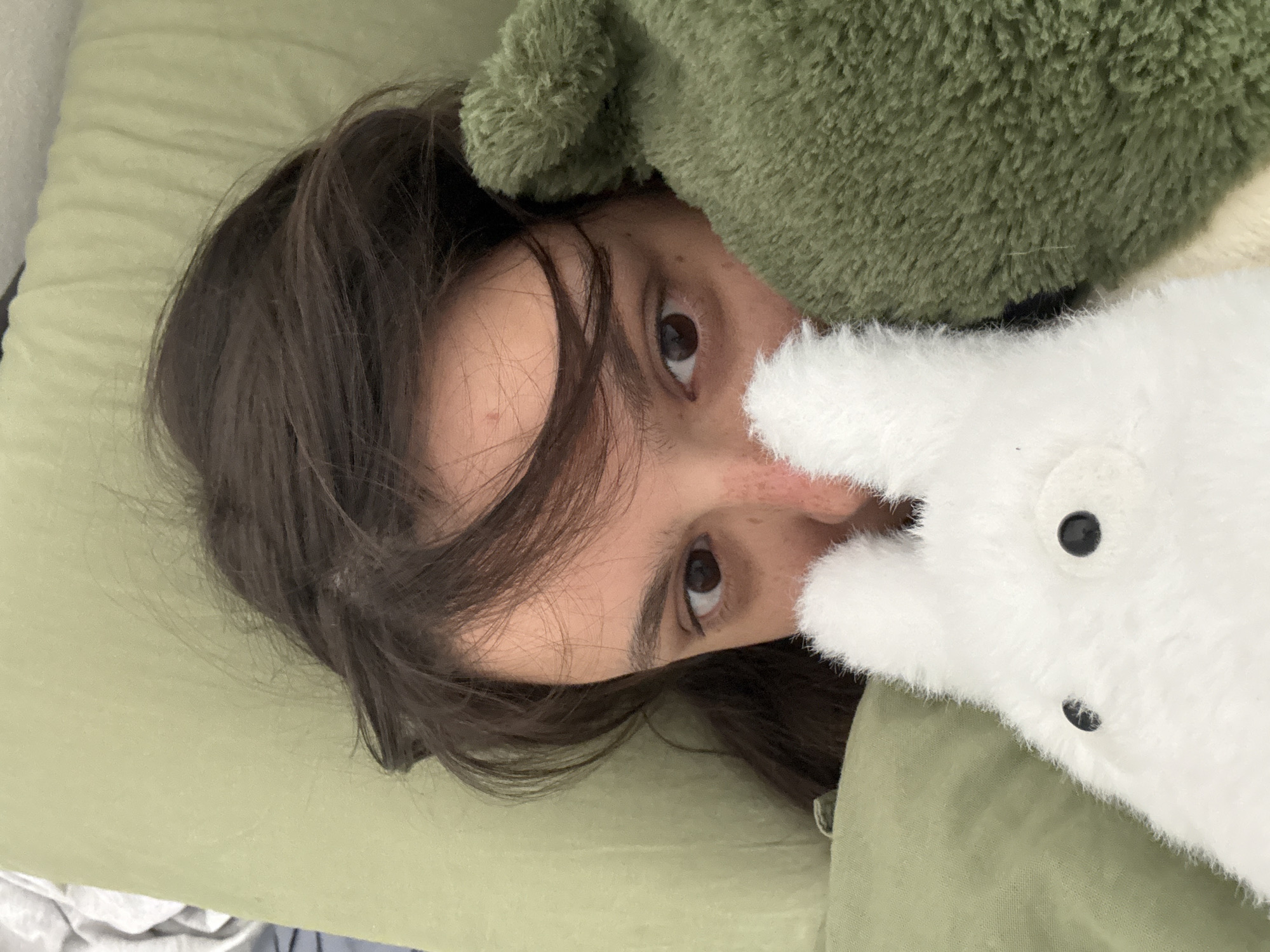 A woman with dark hair and brown eyes is lying on a green pillow and covered with a green blanket. A green, fuzzy object is above her head, and a white rabbit plush toy is next to her face. The background is out of focus, so there is no indication of the location.