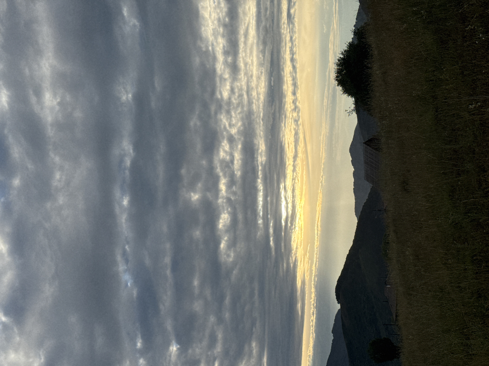 The image depicts a landscape scene with a focus on the sky and rolling hills. The sky is filled with dramatic, textured clouds, primarily in shades of gray and white, with streaks of yellow and bright light suggesting the presence of the sun, possibly at sunrise or sunset. Below the sky, a series of dark, silhouetted hills form the background. In the mid-ground, there is a small, dark-roofed structure, possibly a shed or small house, nestled amongst the hills. The foreground consists of a grassy field, rendered in dark, muted tones, with tall grass and some scattered vegetation. The overall mood is serene and perhaps a bit somber due to the dark color palette and the overcast sky. The specific location within Soloneshnoye, Russia, is not identifiable from the visual information provided.
