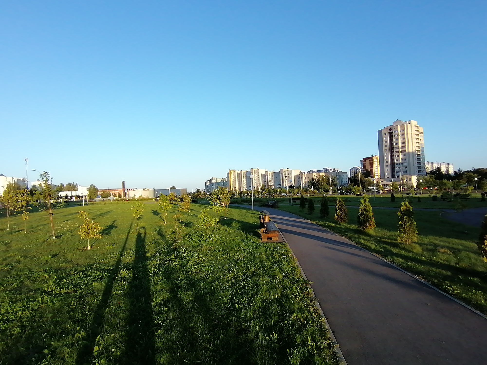 The image shows a park scene in the city of Hrodna, Belarus. In the foreground, a paved pathway curves to the right, bordered by green grass. Several young trees are planted in the grassy area, and two wooden benches are visible along the pathway. Long shadows are cast across the grass, suggesting the sun is low in the sky. In the middle ground, more trees and a grassy expanse are visible, leading to a row of apartment buildings in the background. The sky is clear and blue. The overall impression is that of a well-maintained urban park.
