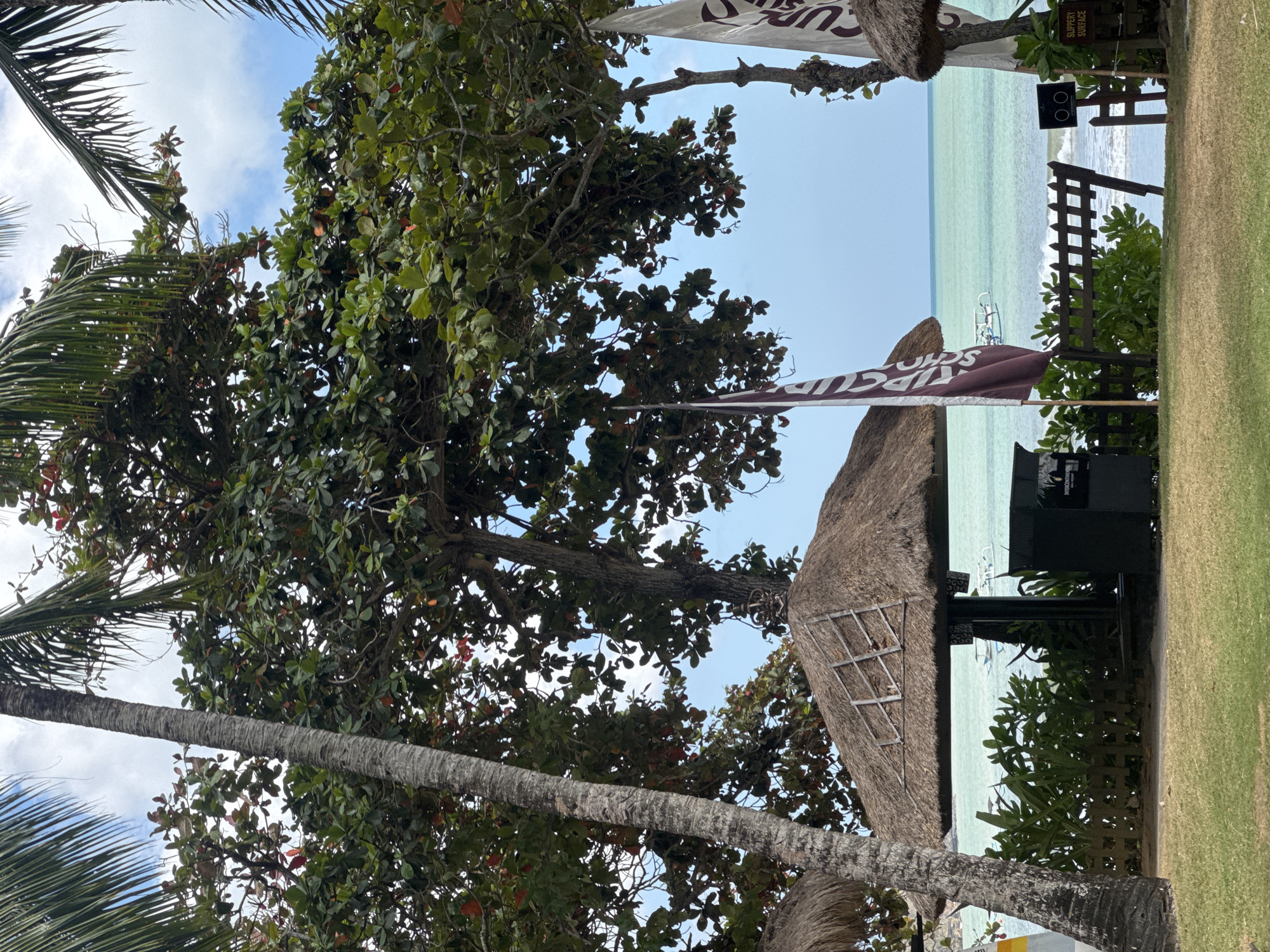 This image depicts a tropical beach scene in Kuta Selatan, Indonesia. The foreground features a neatly trimmed lawn, leading to a pathway or sand area. On the right side of the image, a dark-colored bin with a sign indicating "SLIPPERY SURFACE" is visible. Beyond this, a wooden fence encloses a lush green bush. In the mid-ground, several thatched-roof gazebos or umbrellas are scattered, providing shade. One of these gazebos is prominent, with a dark-colored waste bin underneath it. Two flags are planted near the gazebos; one is a burgundy flag with white lettering, and the other is a white flag with dark lettering.
The background showcases a tranquil ocean with calm, turquoise waters. Small boats, likely traditional Indonesian fishing boats or leisure crafts, are visible on the water. The horizon is clear, meeting a pale blue sky dotted with white clouds. Palm trees and other lush green foliage frame the scene, contributing to the tropical ambiance. The overall impression is one of a peaceful resort or public beach area.