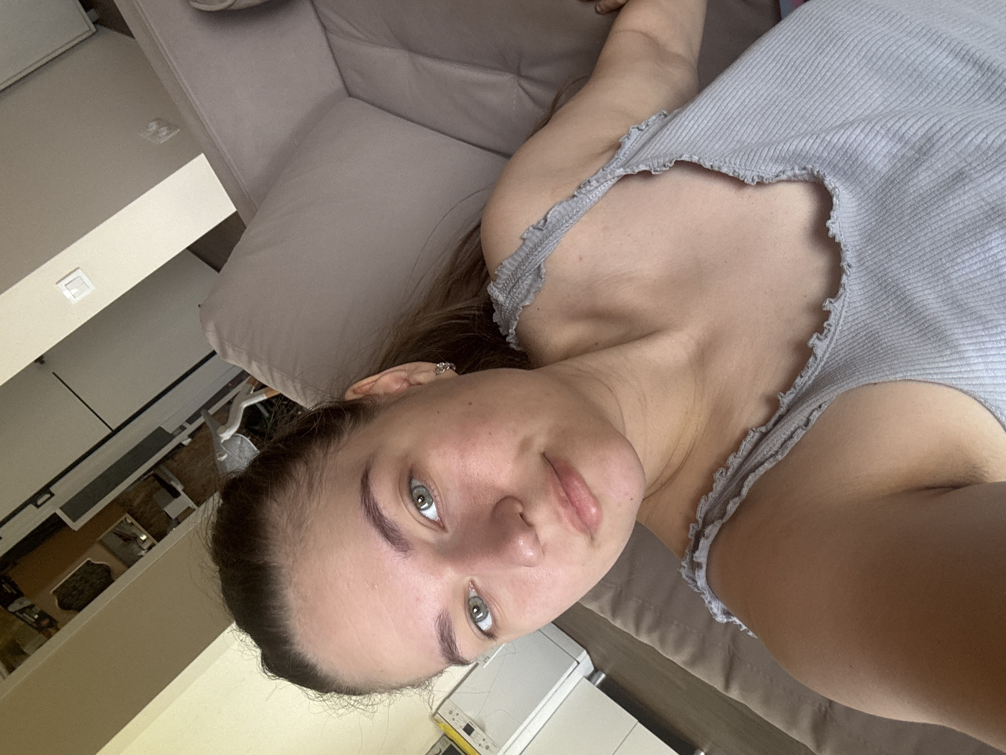 A woman is lying on a light gray couch. She is wearing a light gray ribbed tank top. Her head is oriented towards the lower left of the frame, and her body extends upwards towards the upper right. A cabinet can be seen in the background. It is not possible to determine the location as the city of Chaikovsky, Russia, without additional evidence.