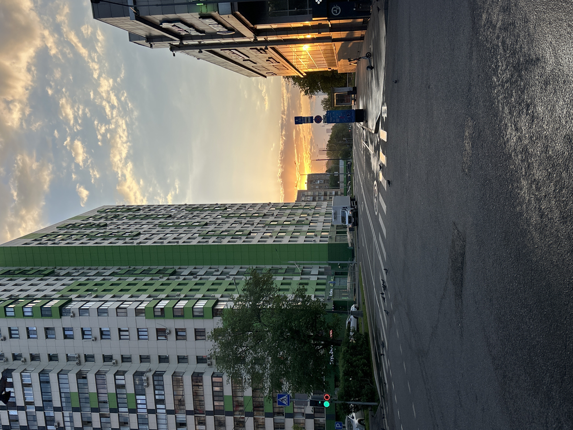 The image shows a street scene at sunset in an unknown city. The sky is filled with clouds and a golden sunset glow is visible between the buildings. On the left is a tall residential building with a pattern of green and white on its facade, and multiple air conditioning units attached. On the right, another building appears to be partially reflecting the setting sun. An asphalt road stretches through the middle of the frame, with white lines marking lanes. A few pigeons are visible on the road. Street signs and a traffic light (showing green) are visible near a tree on the left side of the road.