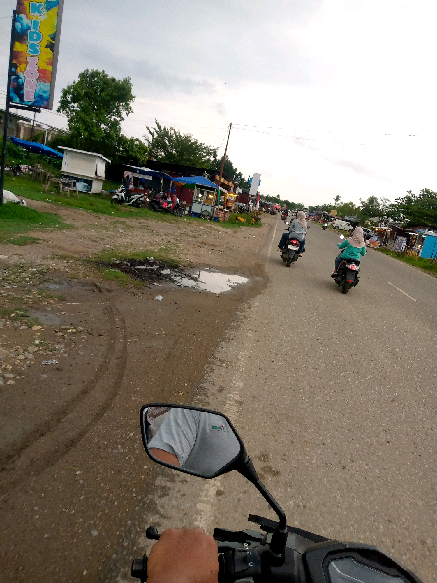 The image captures a scene in Gampongpaya, Indonesia, on a cloudy day, likely in the late afternoon. The perspective is from the point of view of someone riding a motorcycle, with a rearview mirror reflecting the rider's arm and shoulder wearing a gray t-shirt. The road is paved and appears wet in places, with tire tracks visible on the muddy shoulder.
To the left of the road, there are several small stalls and businesses. A tall, colorful sign with the words "KIDS ZONE" is prominently displayed. In front of one stall, a white animal, possibly a goat, is lying down. Several motorcycles are parked near these businesses.
On the road, two women are riding motorcycles in the same direction as the viewer. The woman in front is wearing a light blue shirt and a beige hijab, and the woman behind her is wearing a green long-sleeved shirt and a beige hijab. Both are riding scooters. Further down the road, more vehicles and buildings are visible. The environment suggests a semi-urban or rural setting with roadside commerce.