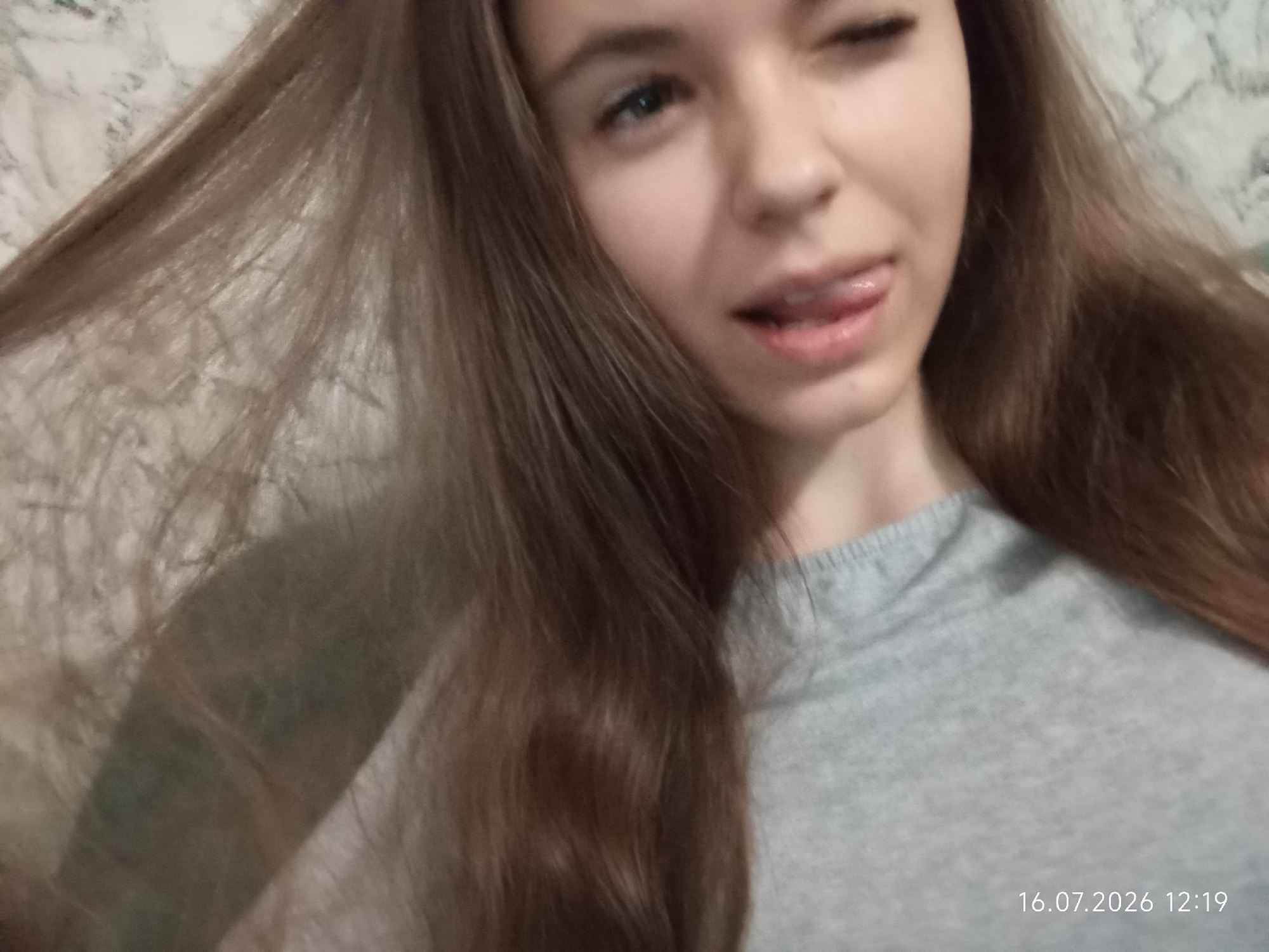 A close-up, selfie-style photograph shows a young woman with long, brown hair. She is winking and sticking her tongue out at the camera. She is wearing a gray t-shirt. The background is blurred and appears to be a wall with a marble-like pattern. A timestamp in the lower right corner reads "16.07.2026 12:19". The image does not provide any information about the city of Qobda, Kazakhstan.