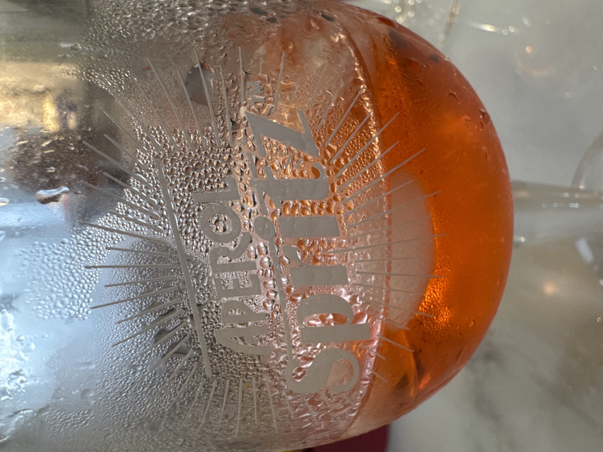 A close-up, waist-up shot shows an Aperol Spritz cocktail in a glass. The glass is decorated with engraved sunburst lines and the words "APEROL SPRITZ" etched into it. Condensation droplets cover the exterior of the glass, obscuring some of the etching. The drink itself is a vibrant orange color, with ice cubes visible within. The background is blurred and appears to be an indoor setting, possibly a restaurant or bar. The image does not provide any information about the location in Chuguyevka, Russia.
