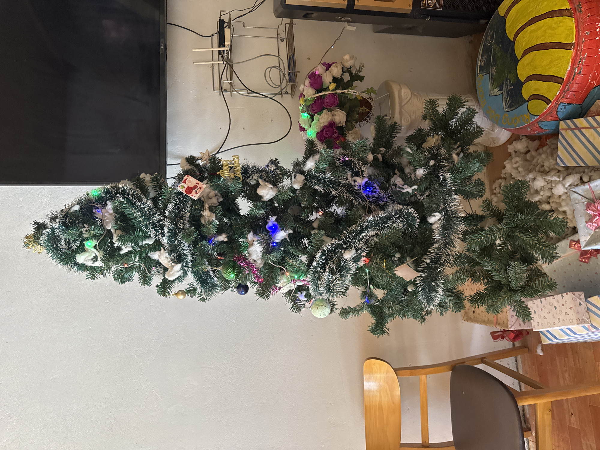A decorated Christmas tree lies on its side in an indoor setting. The tree is adorned with various ornaments, tinsel, lights, and white cotton-like material, suggesting faux snow. Several wrapped gifts surround the base of the tree. Nearby is a bouquet of flowers, a painted tire, a chair, and a television is on the wall. The location is Púng Luông, Vietnam.
