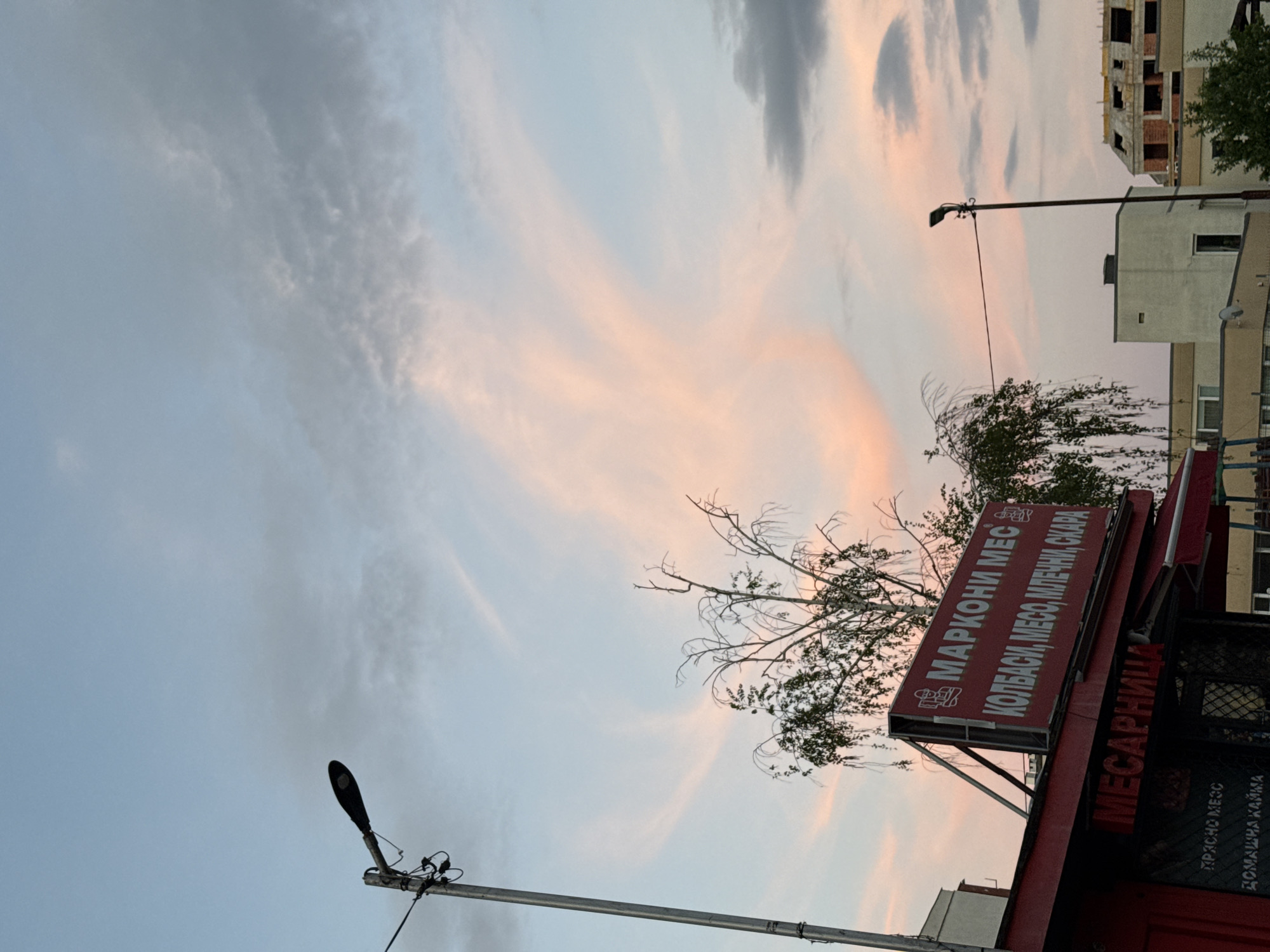 A landscape photograph, initially oriented vertically, captures an urban or suburban street scene at either sunrise or sunset, indicated by the soft, warm light. The sky dominates the upper and central parts of the frame, showcasing a blend of light blue-grey and wispy clouds painted with delicate pink and orange hues. Darker grey clouds are also visible, adding texture to the expansive sky.
In the lower right portion of the image, a prominent red commercial sign belongs to a building, likely a butcher shop or a restaurant specializing in meat products. The sign displays Cyrillic text: "МАРКОНИ МЕС" (Marconi Mes), followed by a list of offerings: "КОЛБАСИ, МЕСО, МЛЕЧНИ, СКАРА" (Kolbasi, Meso, Mlechni, Skara), which translates to "Sausages, Meat, Dairy, Grill." Another part of the sign vertically reads "МЕСАРНИЦА" (Mesarnitsa), meaning "Butcher Shop." Further text on the sign includes "ПРЪСНО МЕСО" (Prasno Meso - "Fresh Meat") and "ДОМАШНА КАЙМА" (Domashna Kayma - "Homemade Minced Meat").
Above and around the sign, the bare branches of a tree reach towards the sky, with some green foliage visible behind it. Another tree with denser green leaves is present further to the right. A modern black streetlight with visible wiring extends from a pole in the middle-left of the image, silhouetted against the sky. Another similar streetlight is partially visible towards the bottom left.
In the background, several buildings are visible. One appears to be an unfinished or under-construction structure with exposed brick and concrete, while others are completed buildings, possibly residential or commercial, adding to the urban setting. The overall scene conveys a quiet moment in a city environment during the soft light of dawn or dusk. The location is Sofia, Bulgaria.