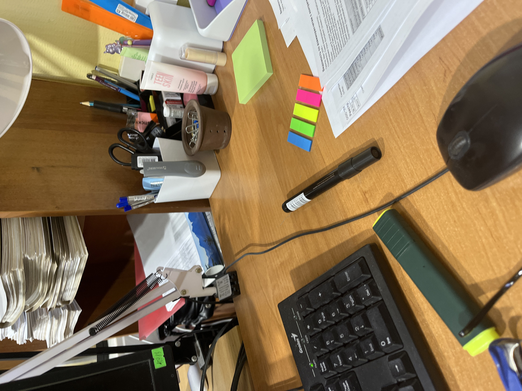 The image depicts a cluttered desk, likely indoors. The scene is dominated by office supplies and paperwork. A black computer keyboard and mouse are visible on the right side of the frame. In the upper-left corner, there is a lamp and an organizer filled with pens, pencils, and a tube of "TEEN SKIN" lotion. A brown container holding paper clips is also in this area. Scattered across the desk are documents with Russian text, along with sticky notes and highlighters. The text on one of the papers mentions parameters related to "Reestr," "Kontragent," "Magazin," and "Dokument." A black marker is lying near the keyboard. The location is possibly in Izhevsk, Russia, based on the Cyrillic script in the documents.