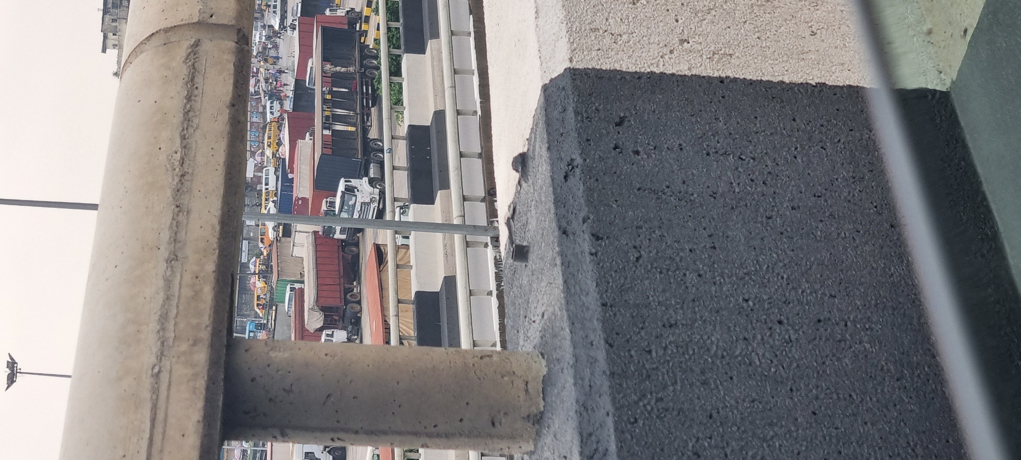 The image captures a busy street scene in Alagbede, Nigeria, as viewed from an elevated position behind a concrete barrier. In the foreground, a large, light-colored concrete pipe runs horizontally across the frame, obscuring a portion of the scene below. Below this pipe, a collection of various trucks and smaller vehicles are visible, suggesting a commercial or transportation hub.
Several large trucks, some with trailers, are parked or moving on a road or lot. One prominent red trailer truck is visible, along with white trucks. In the background, a dense assortment of vehicles, including yellow buses (possibly Danfos, a common form of public transport in Nigeria), cars, and more trucks, indicates significant traffic and activity. The environment appears to be an urban setting, with a slightly overcast sky. The presence of people is suggested by the various vehicles, implying a populated and active area. The overall impression is of a bustling, somewhat chaotic, traffic scene in a Nigerian city.