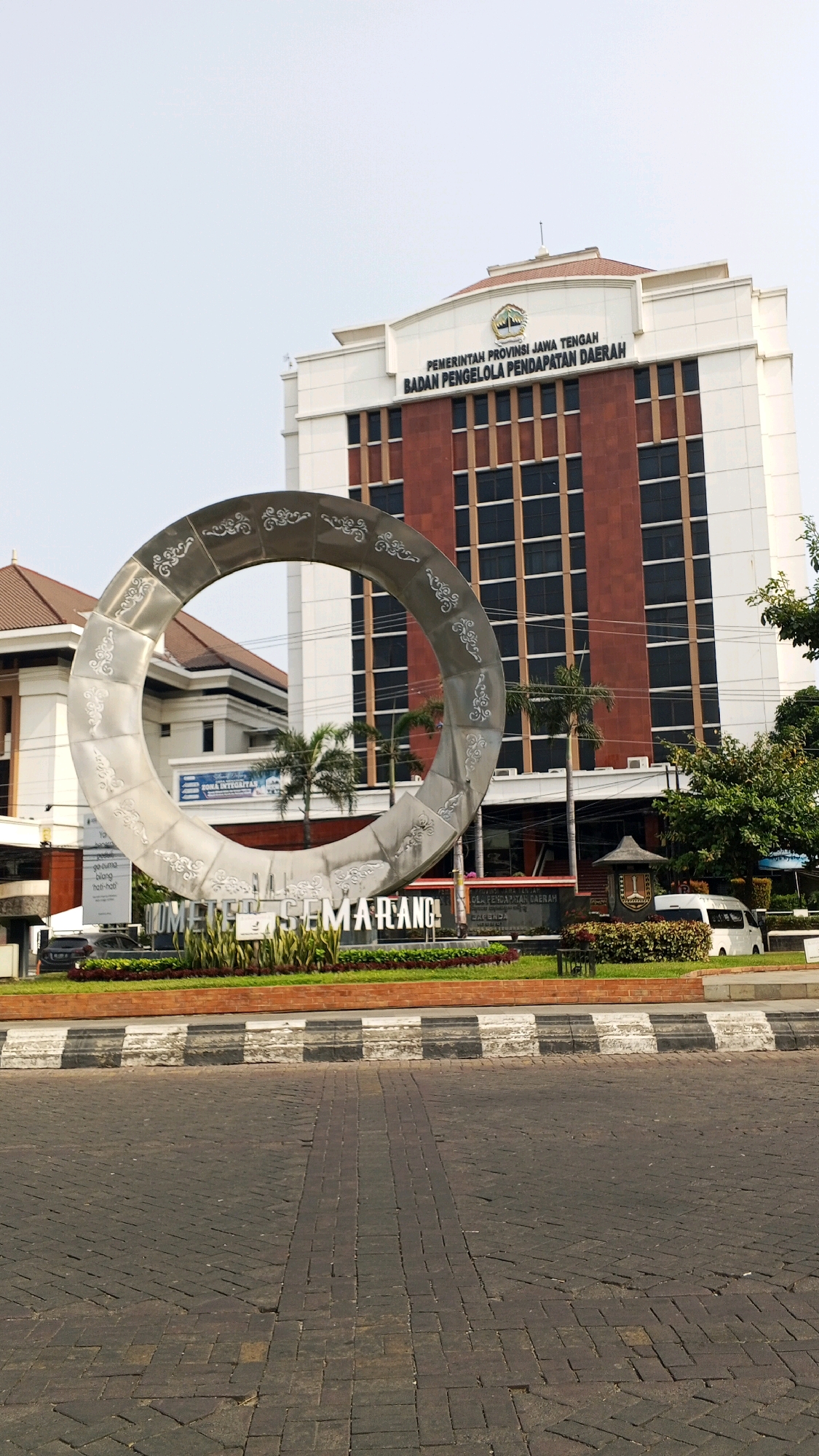 This is a medium shot, eye-level, vertical photograph of the Tugu Muda roundabout in Semarang, Indonesia. The prominent circular sculpture, a significant landmark, dominates the foreground. The sculpture is metallic, silver-toned, and adorned with intricate, swirling patterns. Behind it, a modern, multi-story building with a reddish-brown facade and white trim is visible. Text on the building reads "PEMERINTAH PROVINSI JAWA TENGAH BADAN PENGELOLA PENDAPATAN DAERAH." A white van is parked near the entrance of the building. In front of the sculpture, landscaping with greenery and red-trimmed garden beds is present. The ground in the foreground is paved with dark cobblestones, and a decorative curb with black and white stripes separates the road from the landscaped area. The sky is clear and pale blue. No people are visible in the frame.