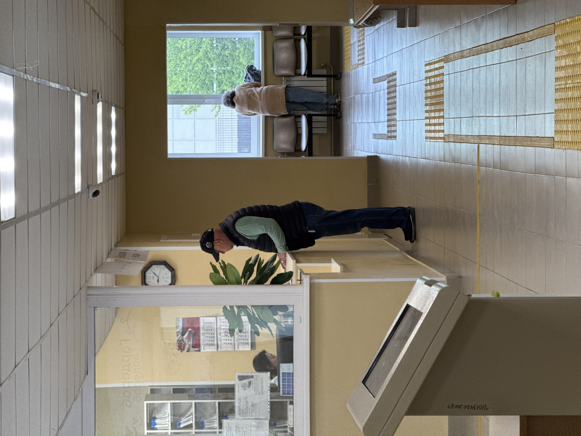 The photograph depicts an interior scene. In the foreground, a middle-aged man is doing push-ups on a window counter. He is wearing a cap, a light green shirt, a dark vest, jeans, and black shoes. His reflection is visible in the window, and a plant sits atop the counter next to him. A digital payment terminal sits at the edge of the frame. A clock on the wall displays the time as approximately 5:55. In the background, a woman is seen sitting and looking out of a window. The walls are painted a neutral yellowish-beige color. The floor is tiled. The location is a public indoor space, possibly a clinic or administrative office.